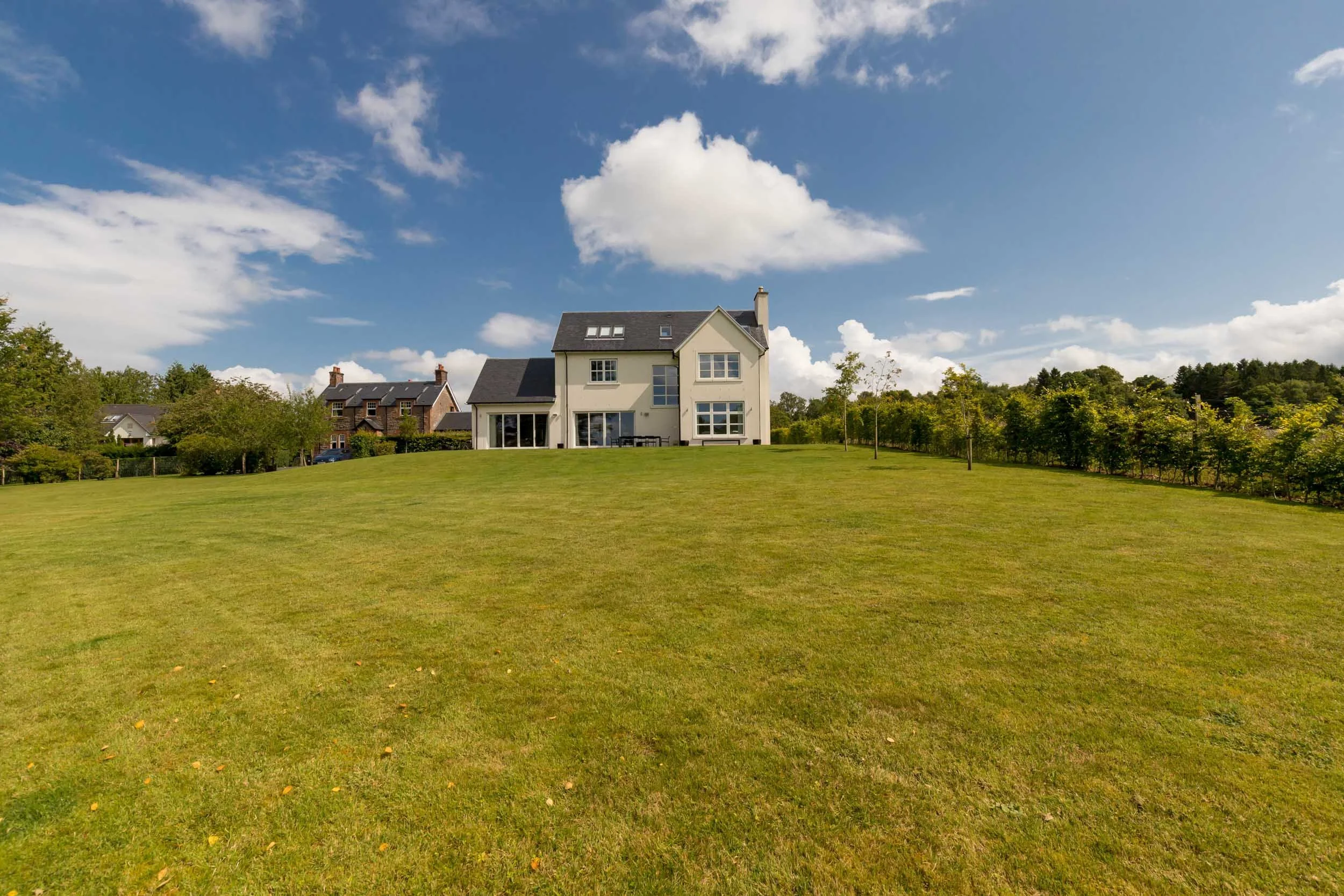 Large white house with multiple windows on a hill under a partly cloudy blue sky, with a well-maintained grassy yard in the foreground and other houses and trees in the background.