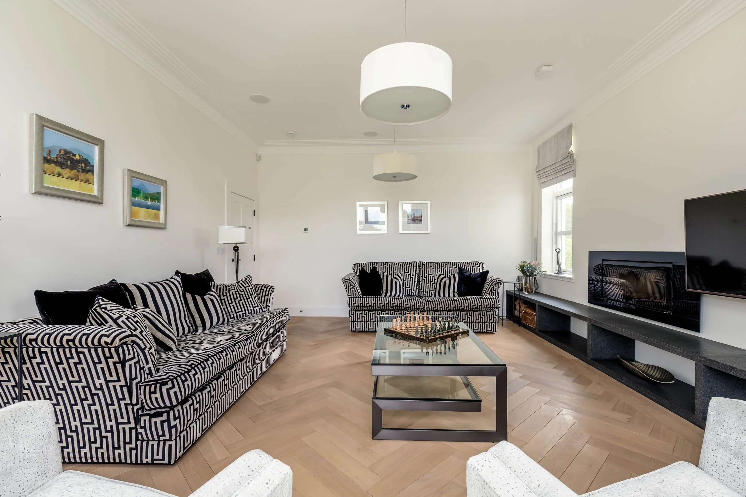 Living room with black and white patterned sofas, a glass coffee table with a chessboard, a wall-mounted TV, artwork on the walls, a window with blinds, and light wood flooring.