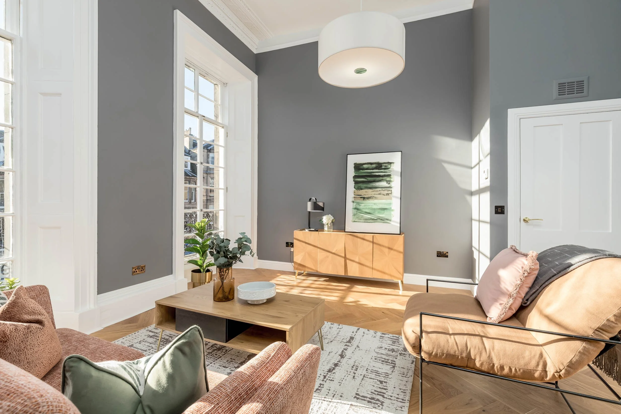 Modern living room with large windows, light wood furniture, gray walls, and colorful cushions.