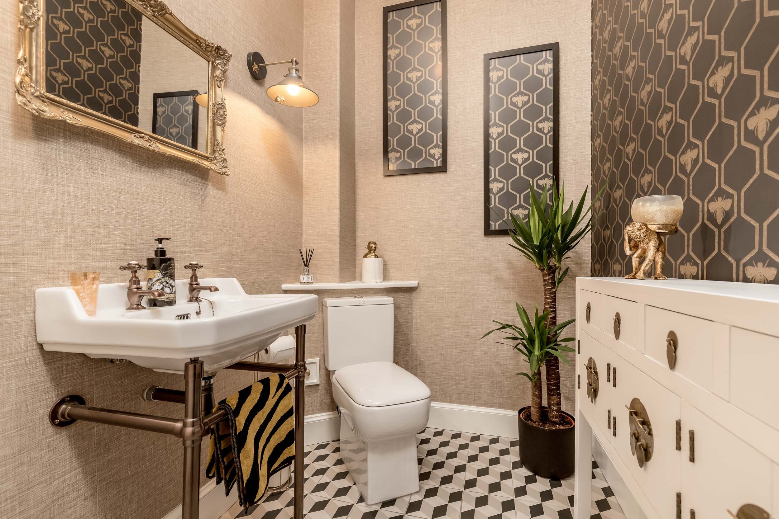 A small bathroom with beige textured walls, a white toilet, and a white vintage-style sink with bronze fixtures. The mirror above the sink has an ornate gold frame, and there is a wall-mounted light fixture beside it. The floor has a black and white 