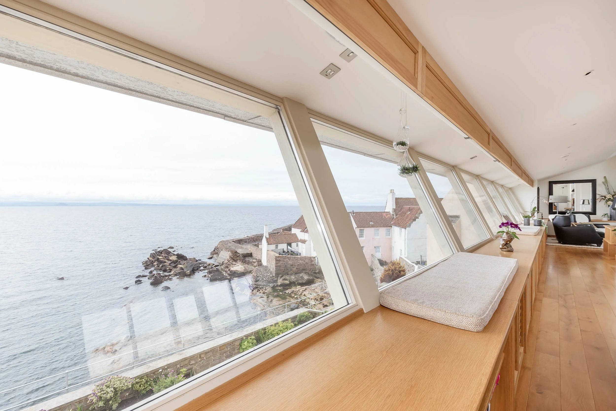 Living room with large front-facing windows overlooking the ocean, with a wooden bench and decorative plants inside.
