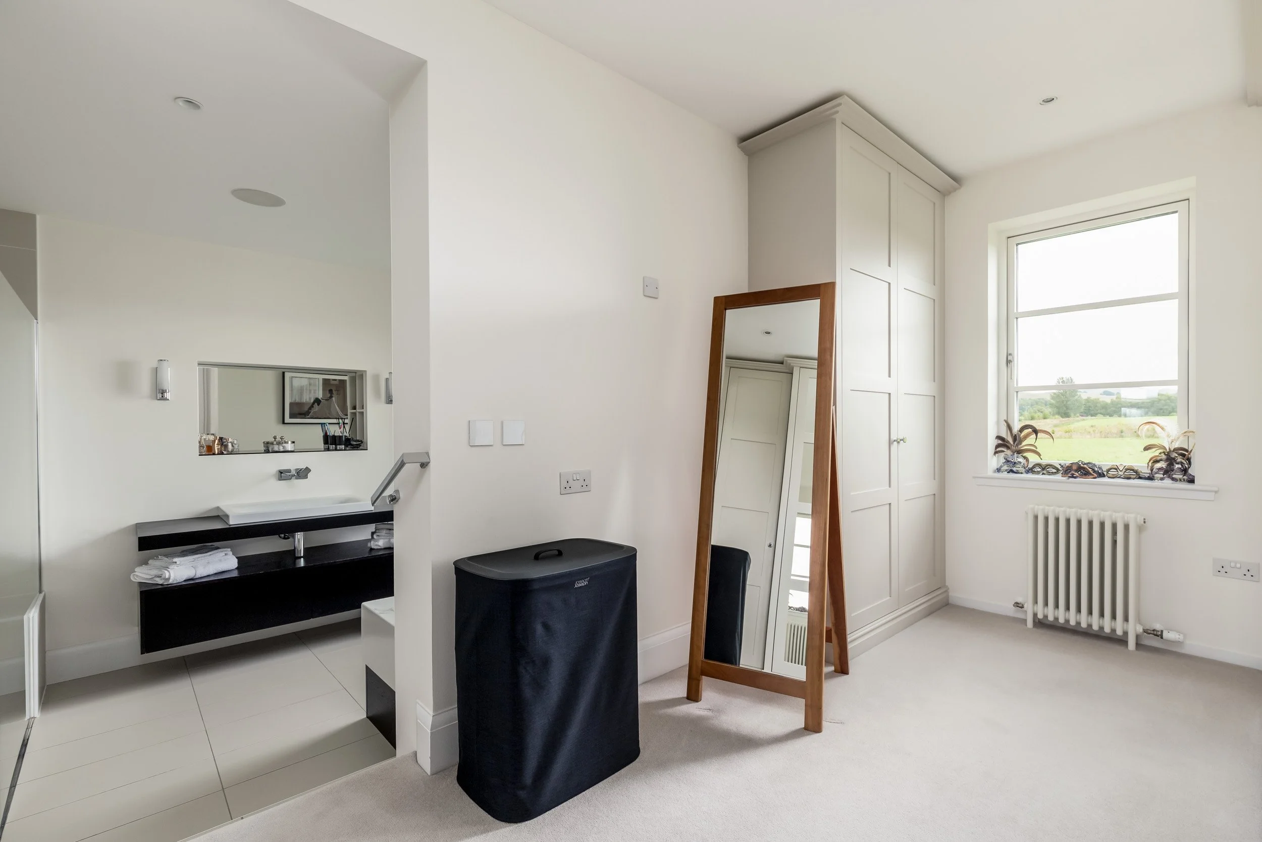 Upside-down mirror leaning against a white wardrobe in a bright room with a window and beige carpet, with a black laundry hamper nearby.