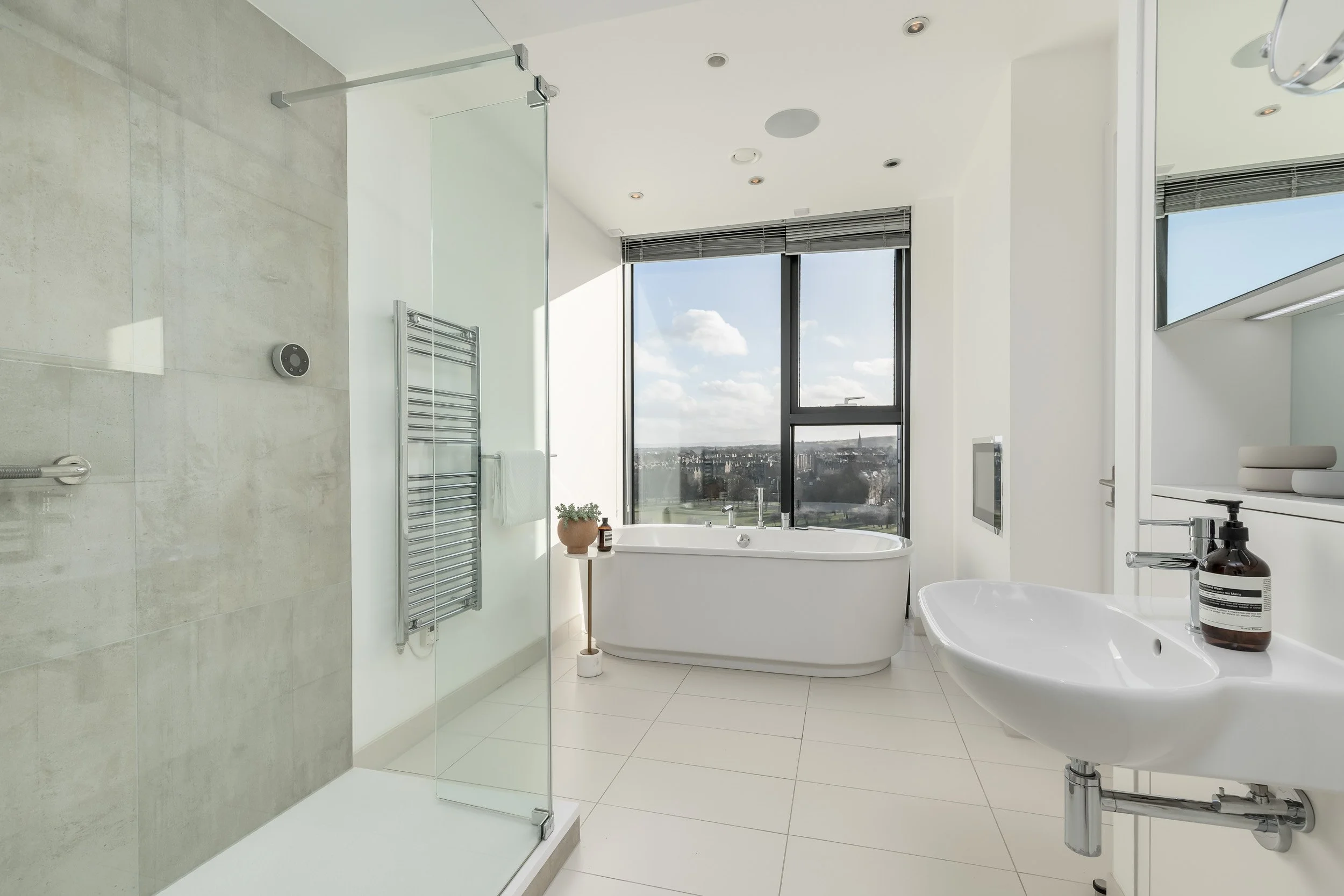 Modern bathroom with a freestanding bathtub, a glass-enclosed shower, a white sink, and a large window view of the city sky.