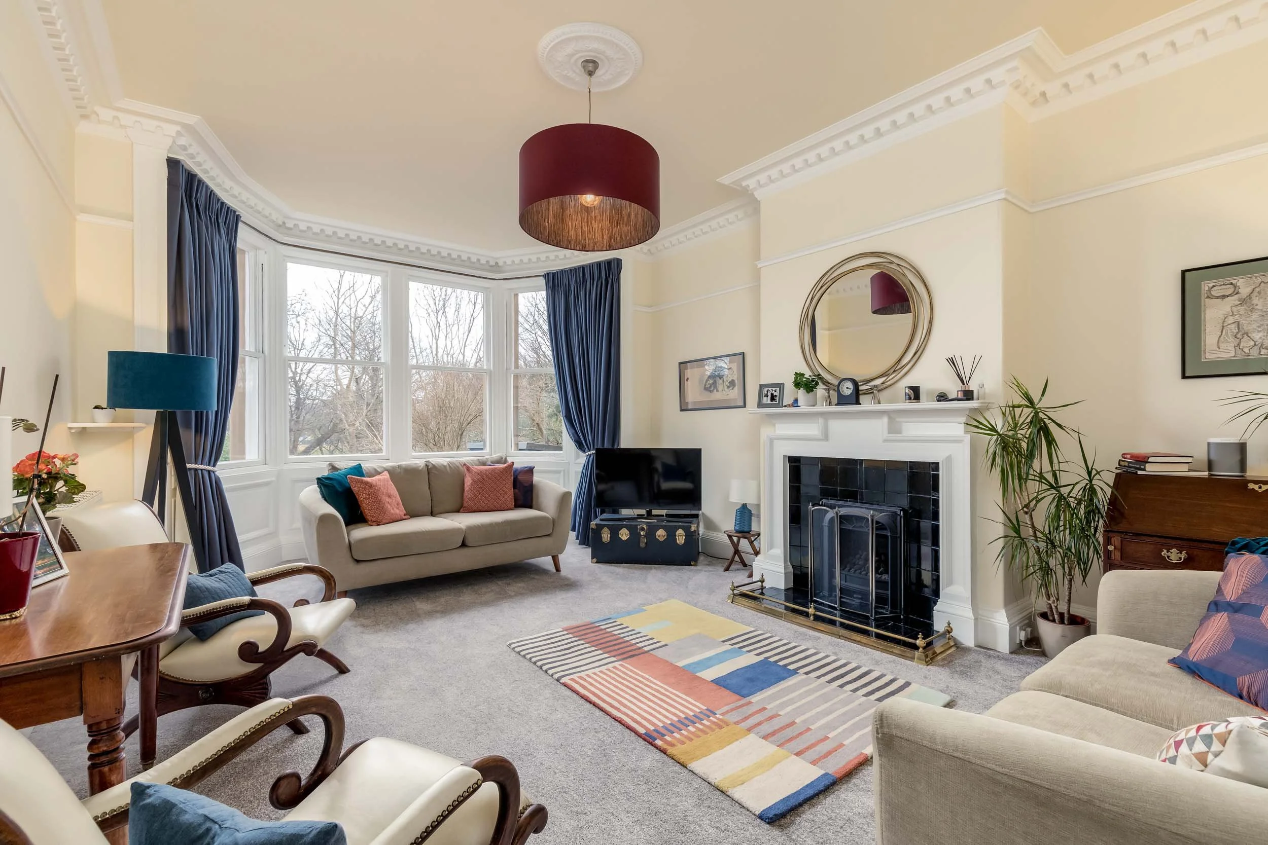 A living room with beige walls, a fireplace with black tiles, a round mirror above the mantel, and a large bay window with blue curtains. The room has two beige sofas with colorful pillows, a colorful striped rug, a TV on a stand, and various decorat