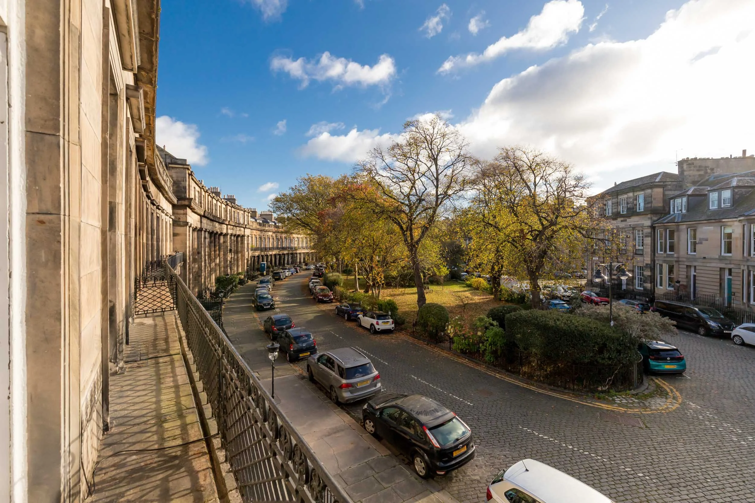 A street scene in a residential neighborhood, with parked cars along the curb, a row of historic townhouses on the left, and a small park with trees and bushes on the right under a blue sky with some clouds.