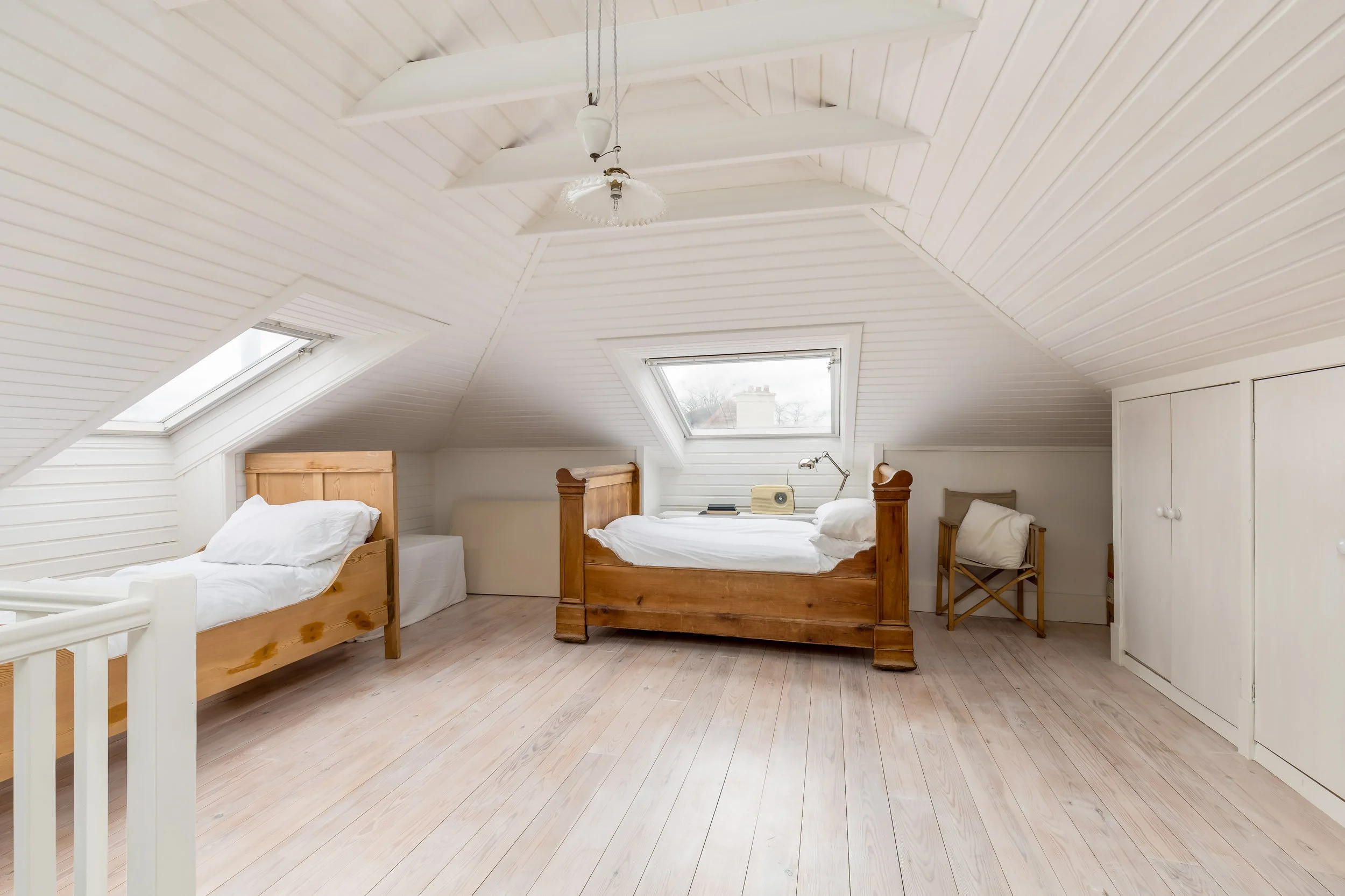 Bright attic bedroom with two wooden beds, white bedding, skylight windows, and light wood flooring.