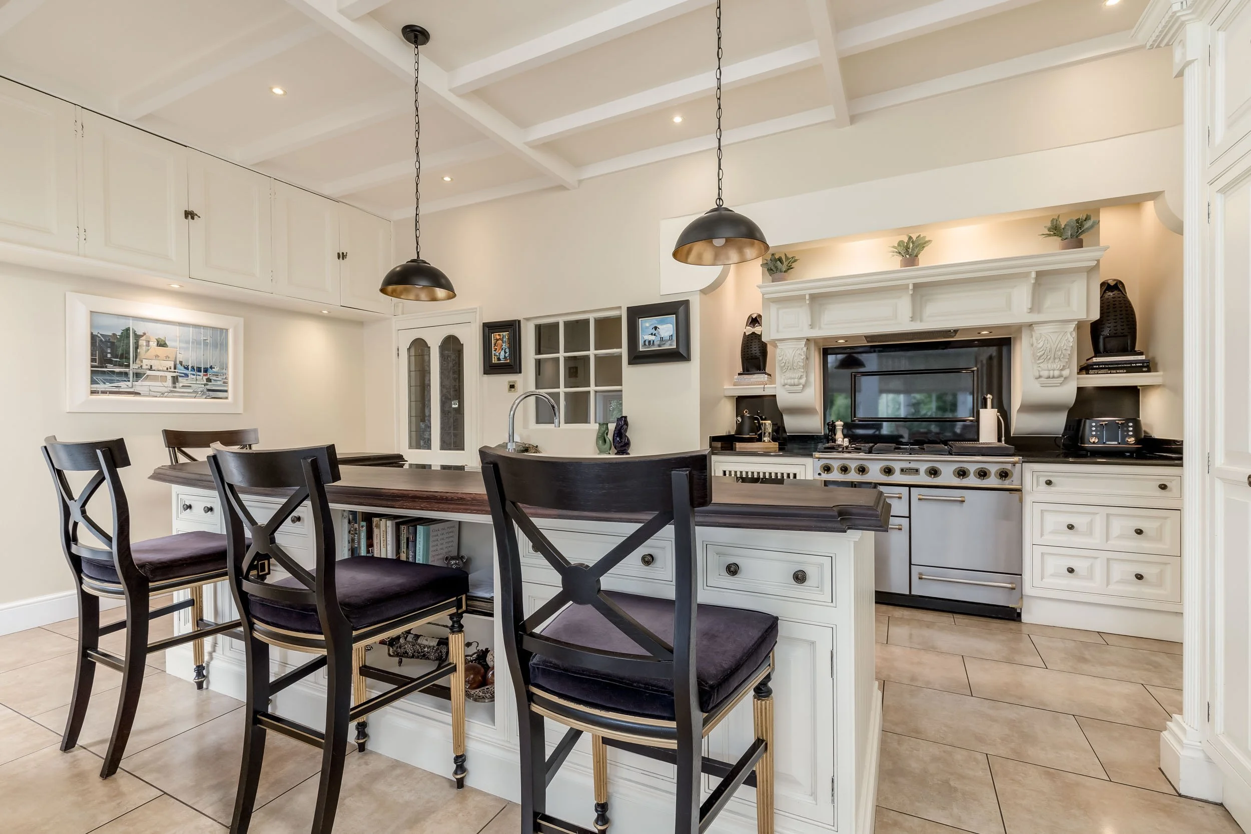 Modern kitchen with white cabinets, a large dark wood island with seating, a stainless steel stove, and decorative plants above the oven. Pendant lights hang over the island, and there is a television above the stove with small decorative items on ei