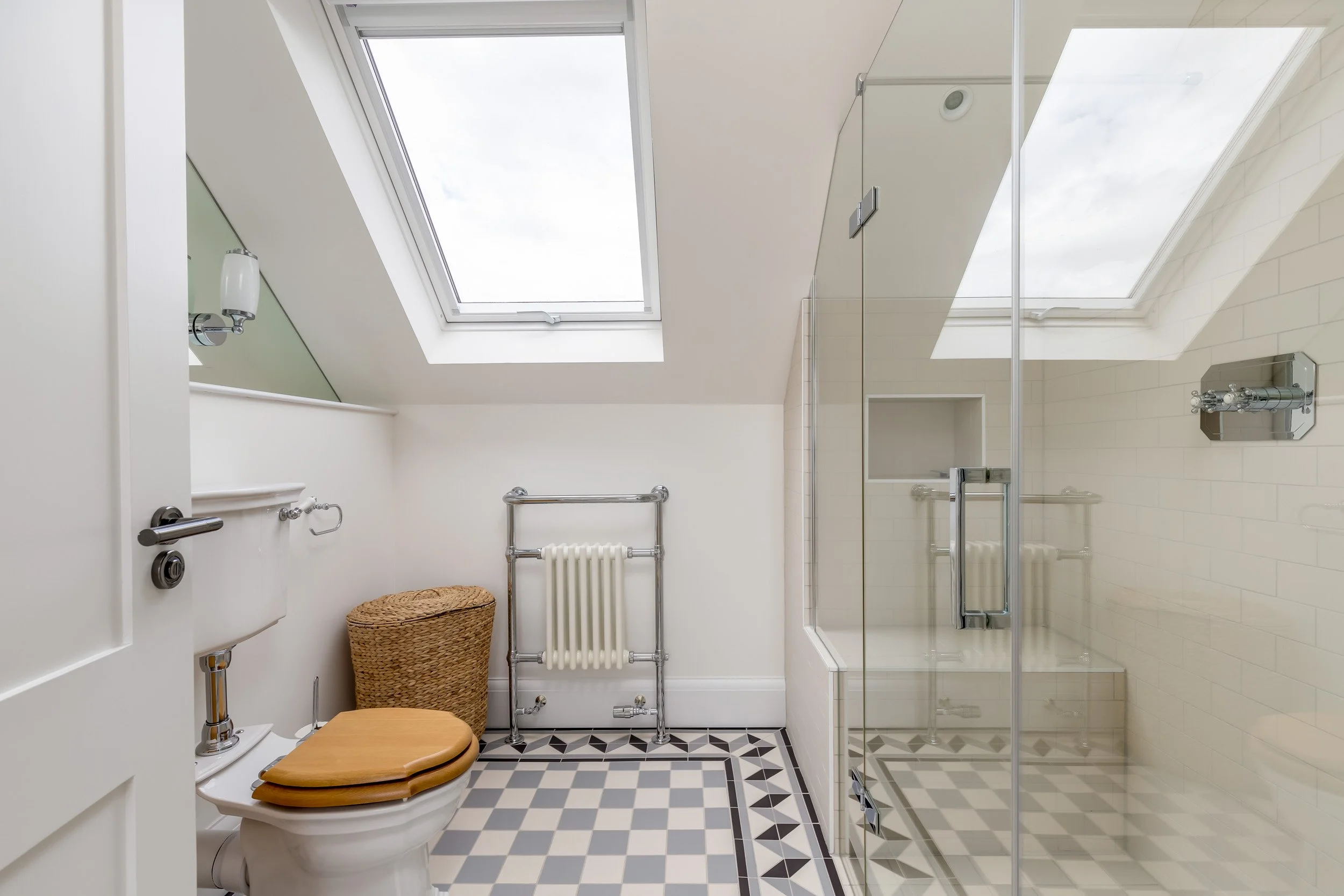 Bright bathroom with a skylight, walk-in shower with glass door, toilet with wooden seat, and a wicker laundry basket.