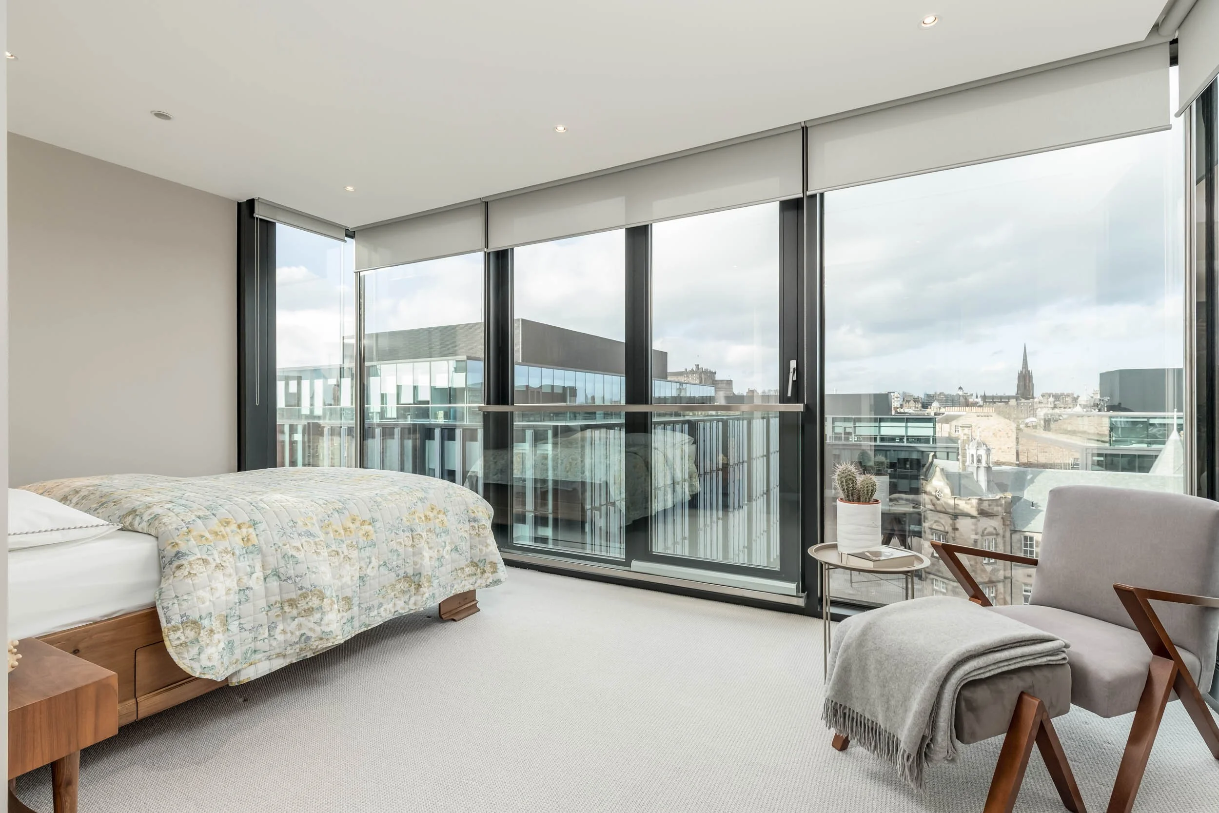 Modern bedroom with large glass windows offering city skyline view, a bed with a floral quilt, a gray armchair with a blanket, and a small side table with a cactus plant.
