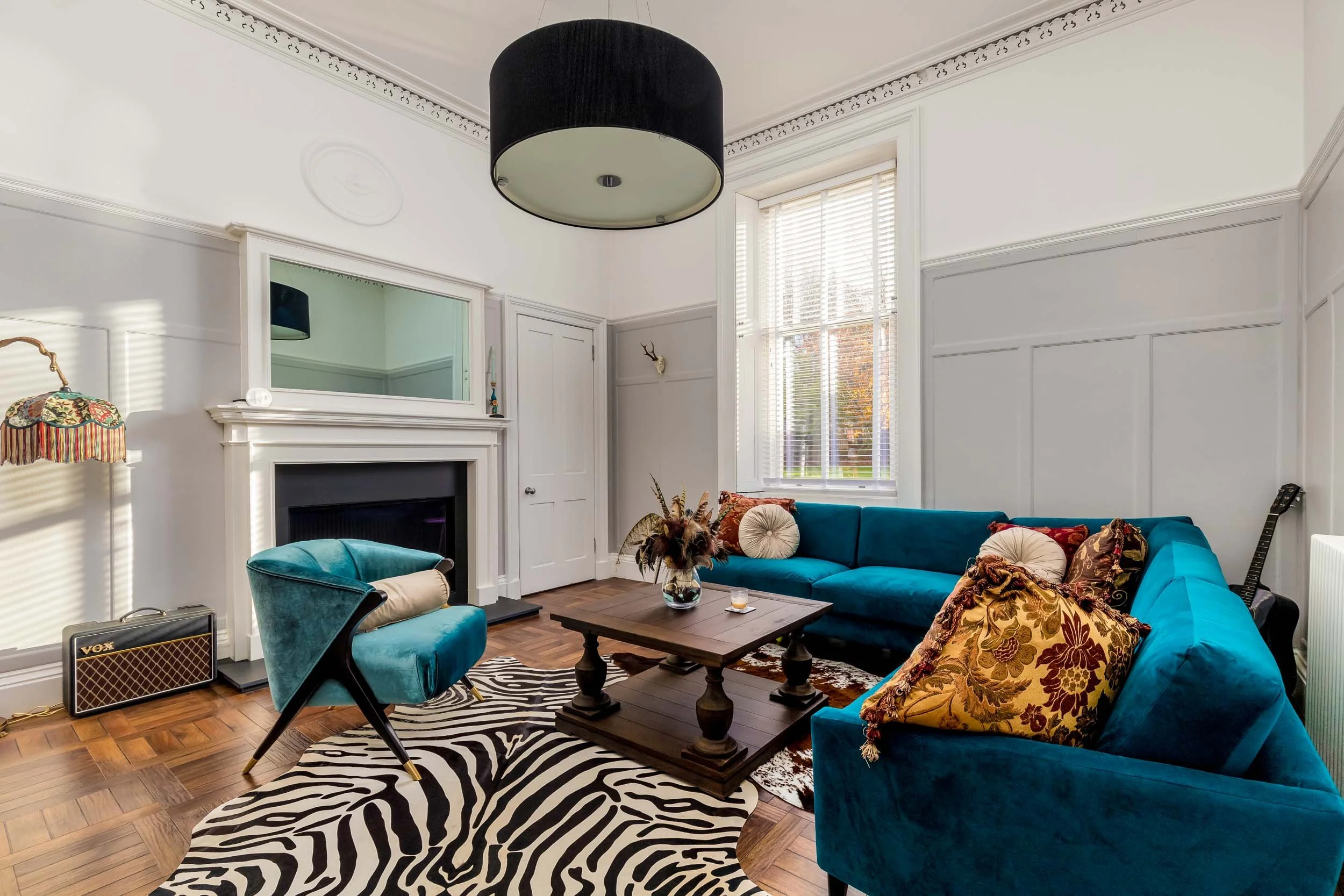 Living room with blue sectional sofa, zebra-patterned rug, wooden coffee table, teal accent chair, fireplace with mirror, window with blinds, and various decorative pillows and items.