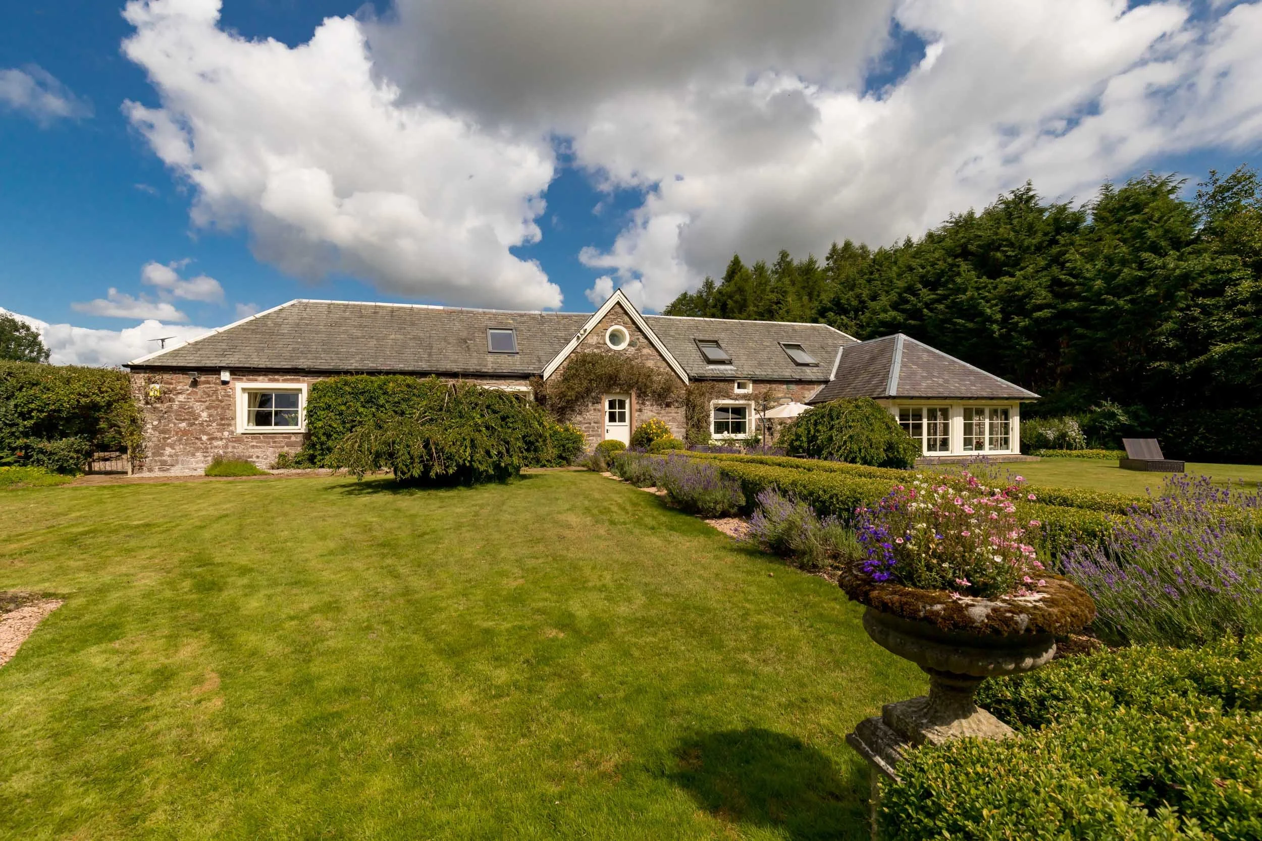 A large, stone and brick country house with a sloped roof set amidst a lush green garden with well-manicured lawn, flowerbeds, and trees. The house has multiple windows, including a small circular window, and a patio area with a white umbrella and ou