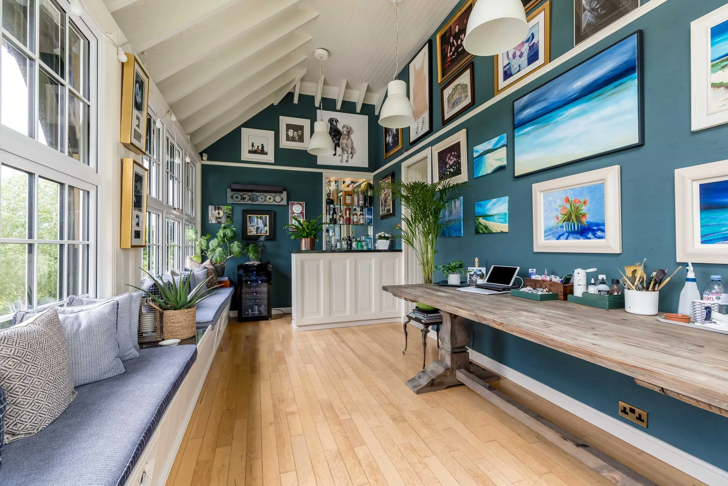 Bright room with teal walls decorated with various framed artwork, a large wooden table with office supplies, a window seat with pillows and plants, and a small bar area with shelves of bottles and plants.