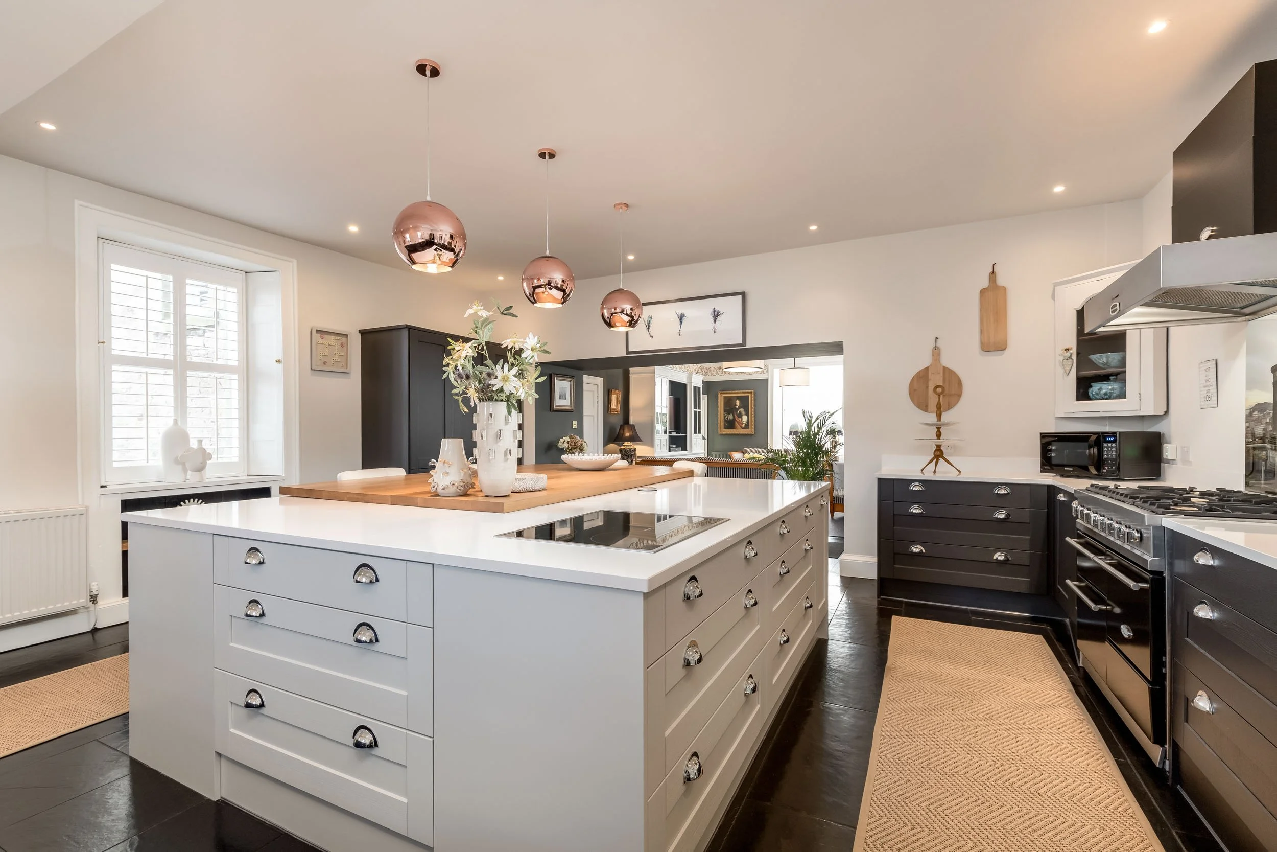 Modern kitchen with white center island featuring silver hardware, black lower cabinets, a black cabinet in the background, and black appliances including oven and microwave. Decorative items and pendant lights hang over the island.