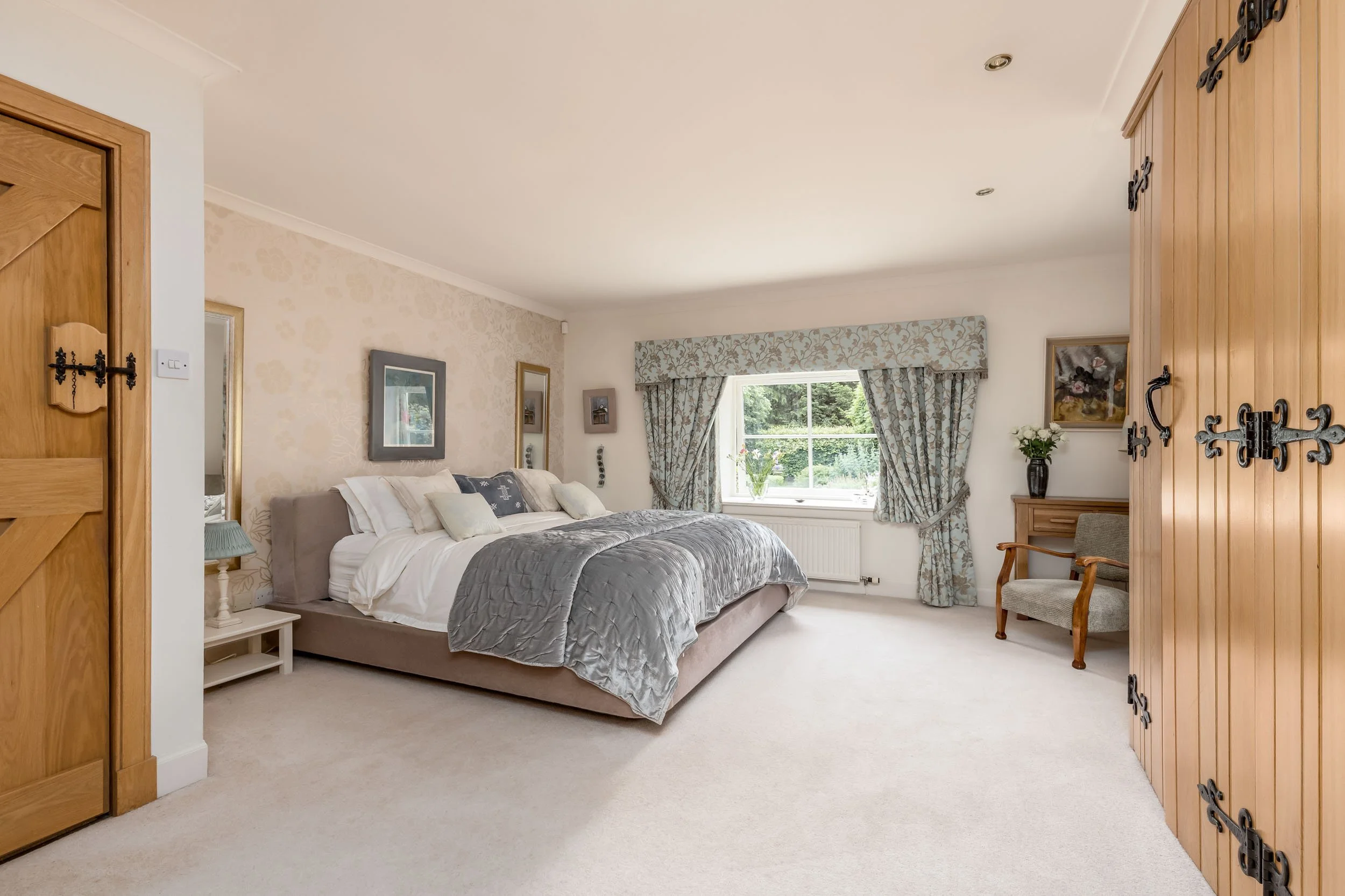 A bedroom with a large bed, floral curtains, a chair, a wooden wardrobe, and framed artwork, with natural light coming from a window.