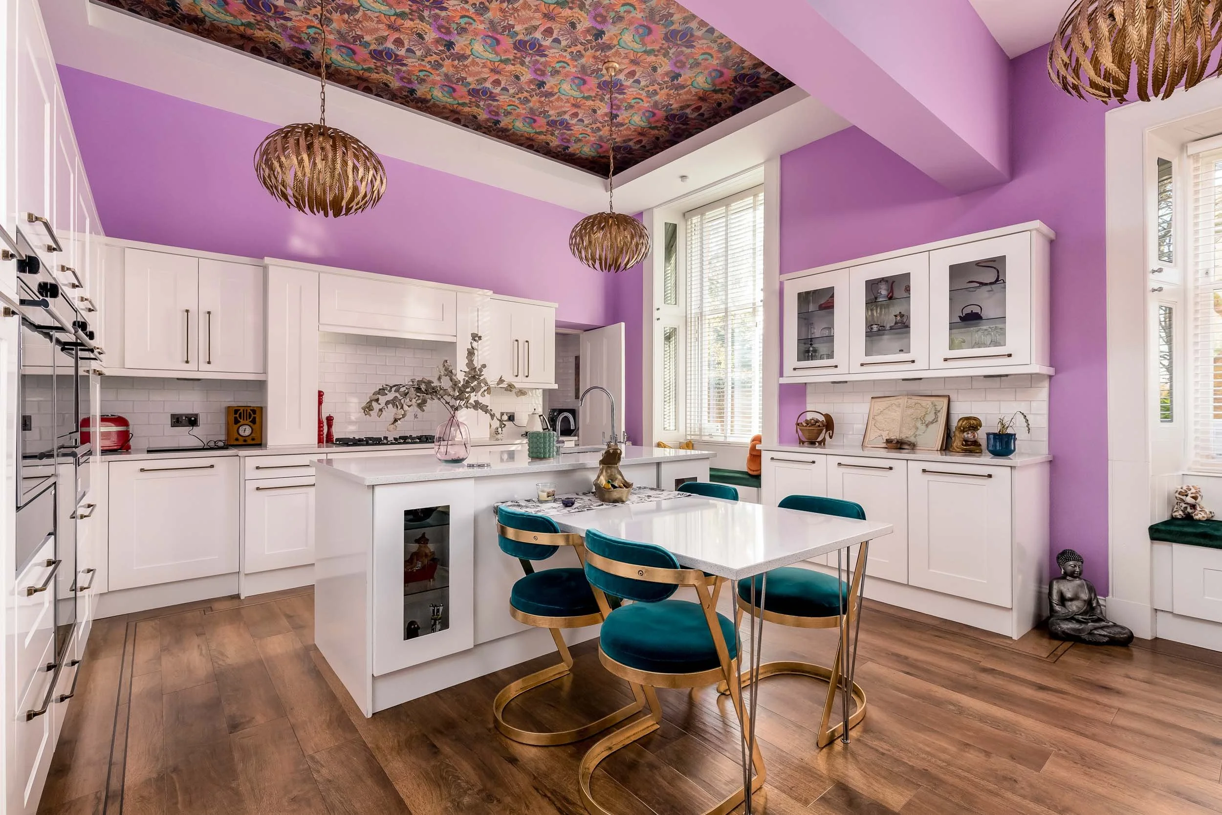 Bright kitchen with purple walls, white cabinets, wooden floors, and a floral ceiling. Contains a kitchen island, teal chairs with gold frames, hanging light fixtures, and decorative items on shelves.
