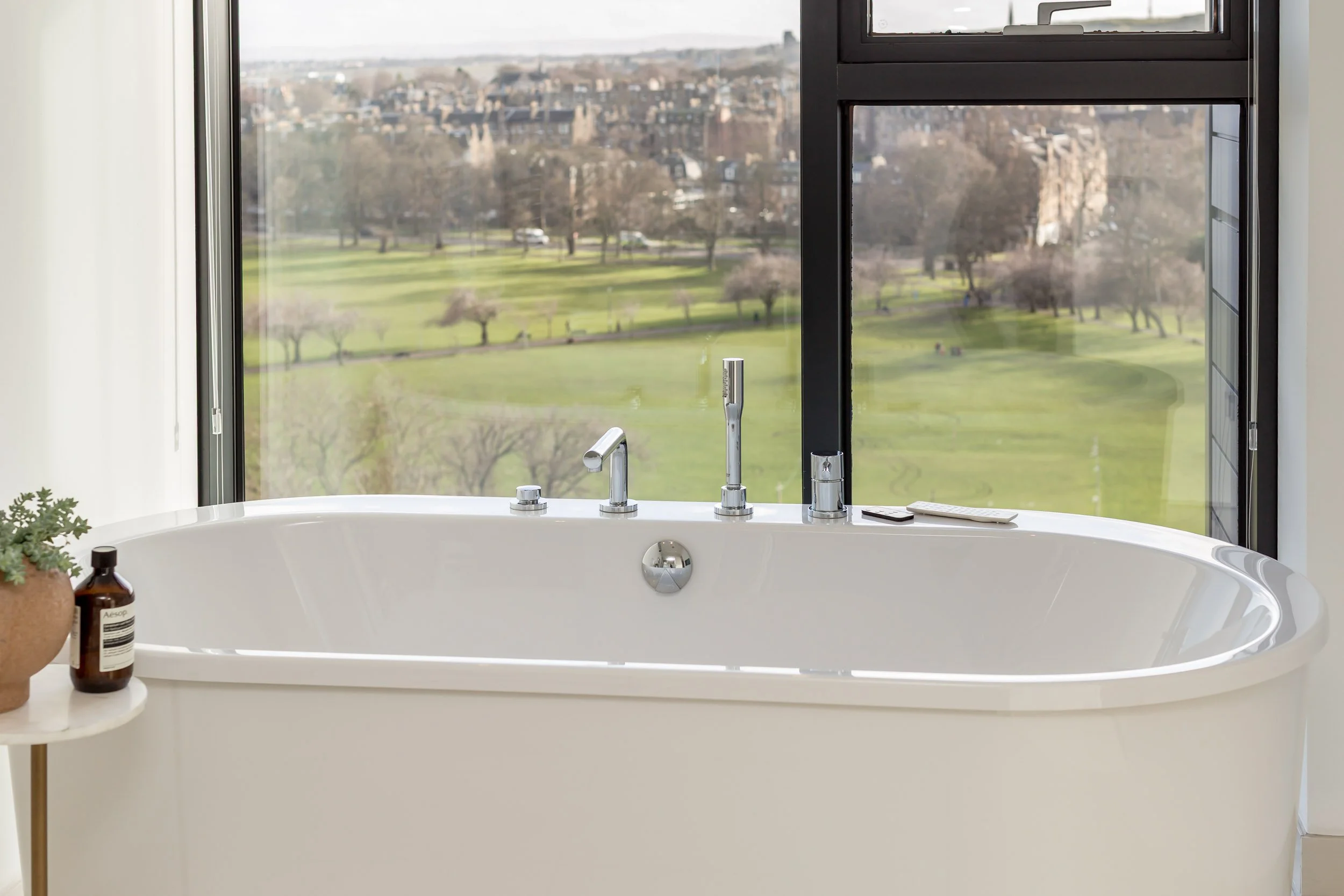Bathroom with a white bathtub positioned in front of large windows overlooking a green park and cityscape in the distance.