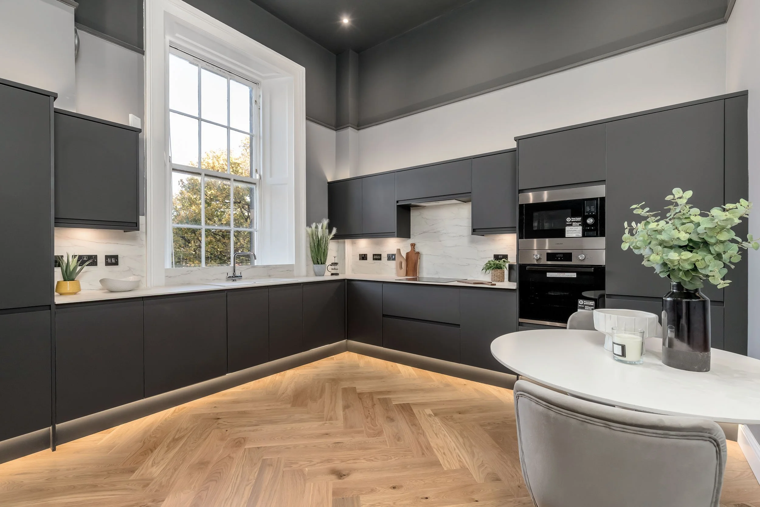 Modern kitchen with black cabinets, white marble countertops, and a large window showing trees outside. There is a small round table with a chair, a flower vase, and decorative candles.