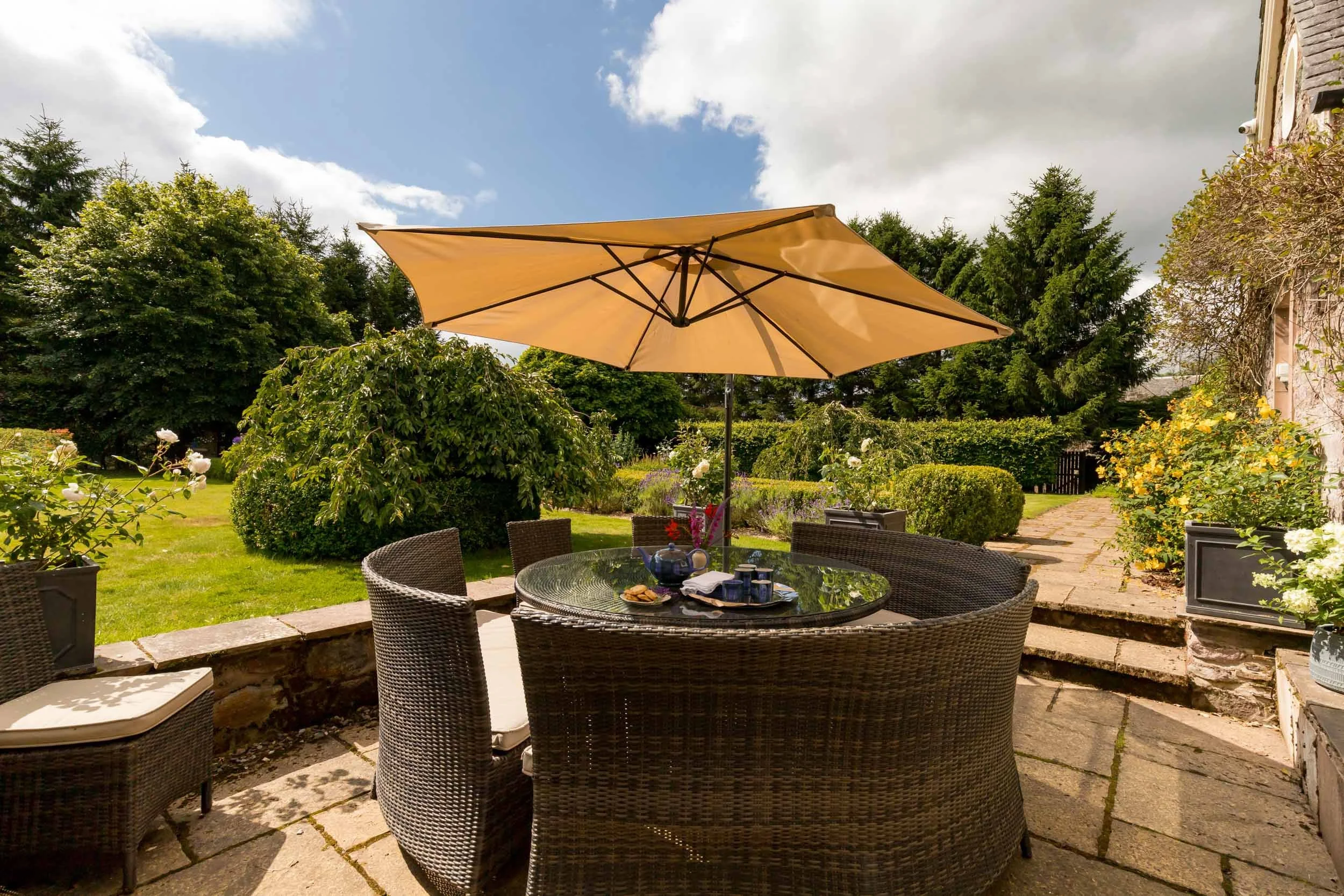 Outdoor patio dining area with a round glass table, wicker chairs, and a large yellow umbrella, set in a garden with bushes, flowers, and trees on a partly cloudy day.