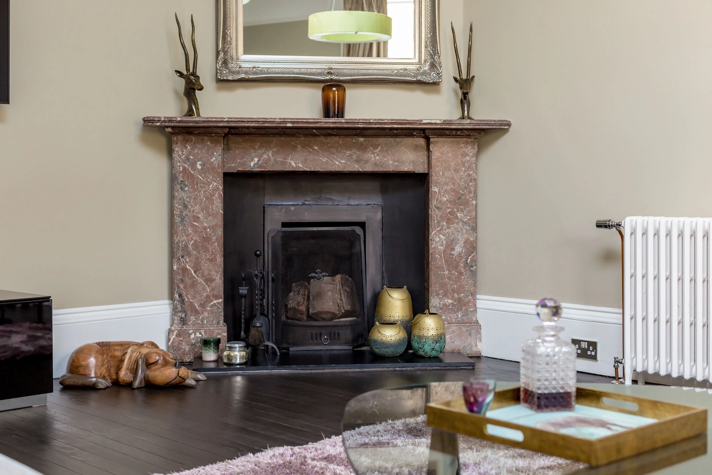 Living room fireplace with a pink marble surround, decorative vases, and golden animal sculptures on the mantel. A mirror hangs above the fireplace, and there's a soft pink rug and a glass decanter with a tray on the coffee table.