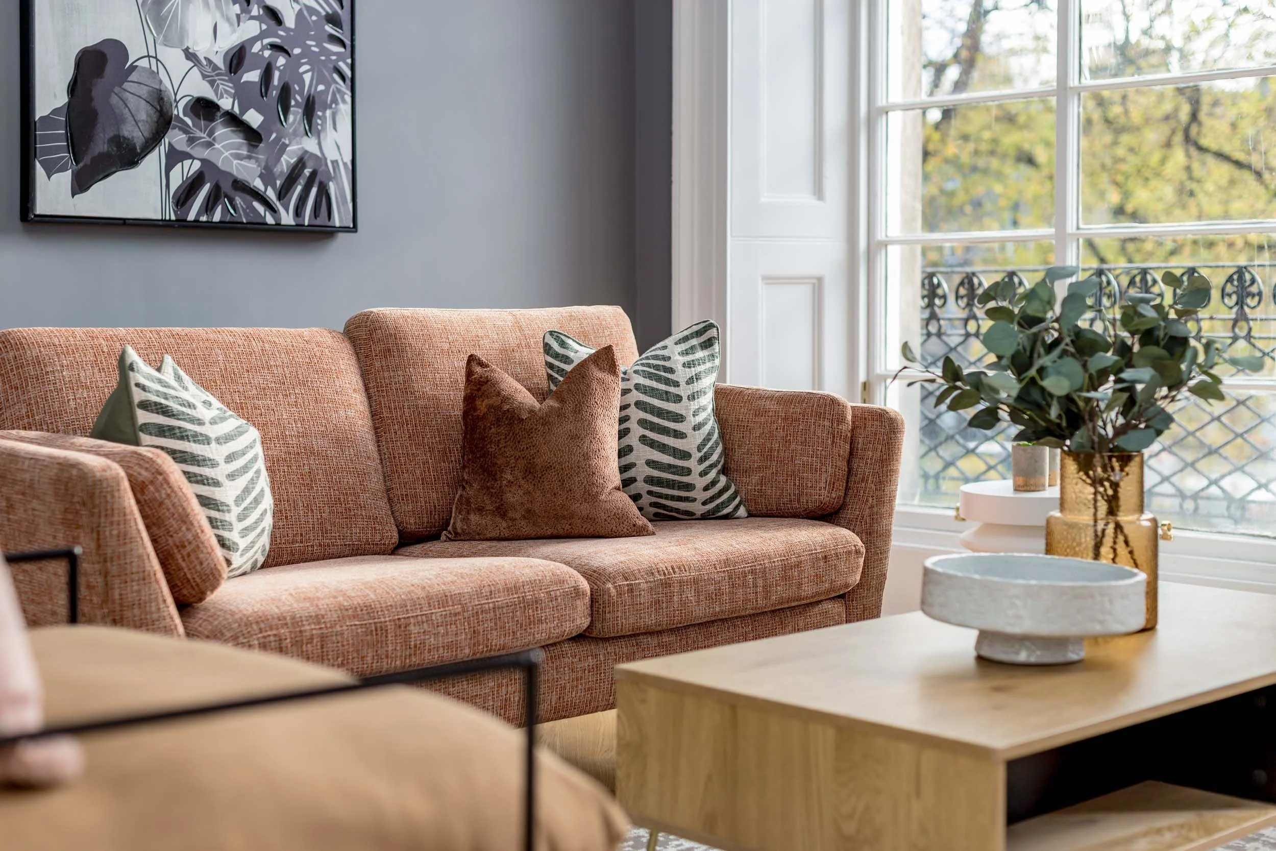 Living room with a pink textured sofa, decorative pillows, a wooden coffee table, a large window with a balcony, a potted plant, and wall art.