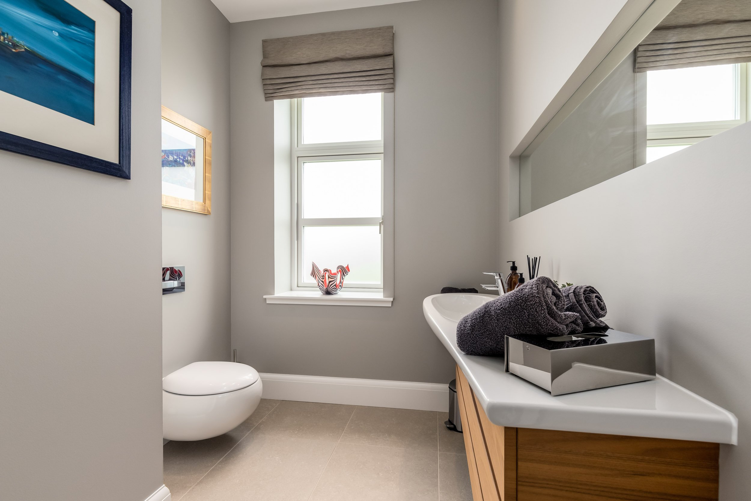A modern bathroom with a white toilet, a window with a beige roman shade, a wooden vanity with a white countertop, a sink, and dark towels rolled on a tray. There are framed pictures on the wall and a decorative vase on the window sill.