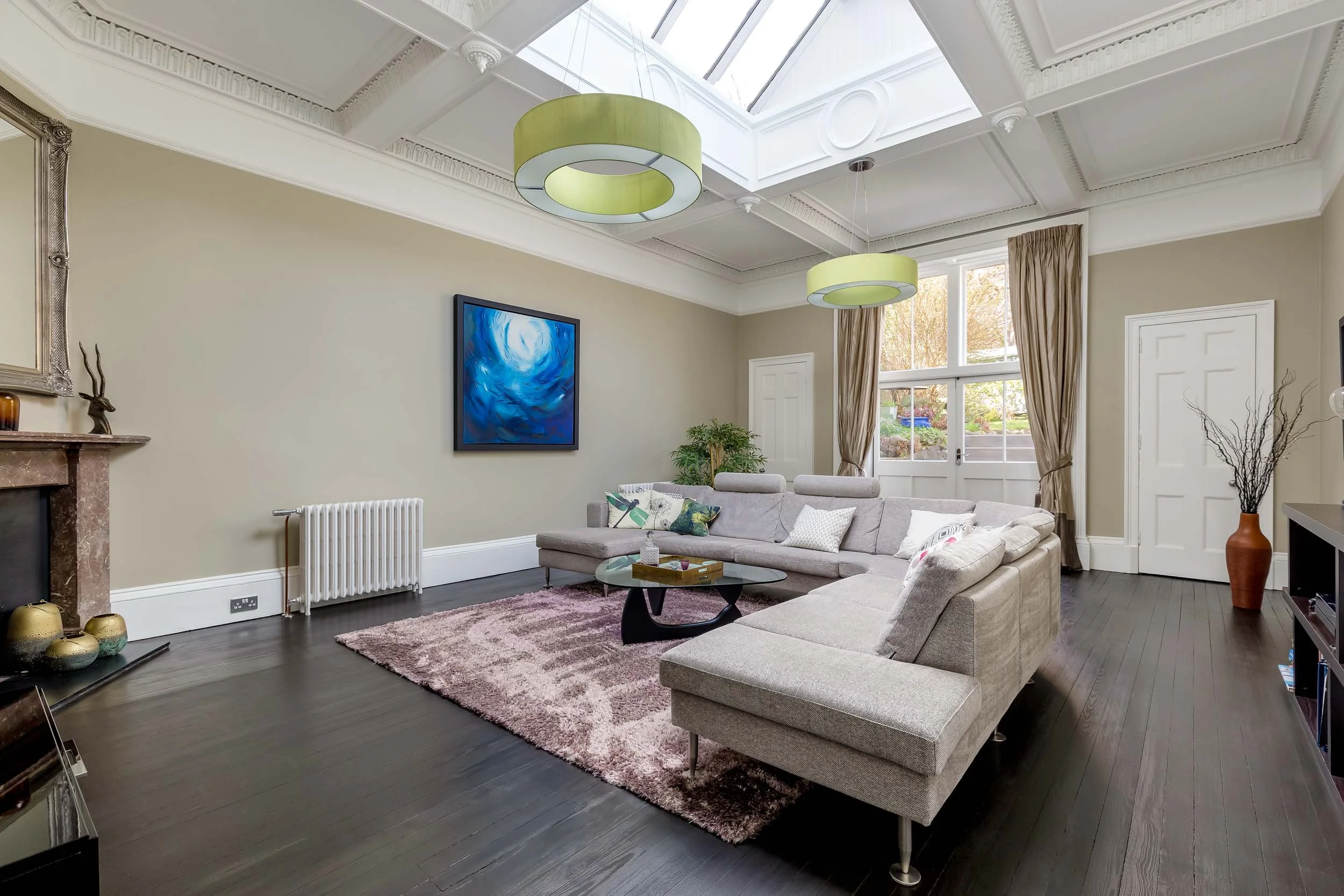 Bright living room with gray sectional sofa, patterned pillows, glass coffee table, dark hardwood floor, beige walls, and large windows with curtains. Modern ceiling lights and colorful wall art are also visible.