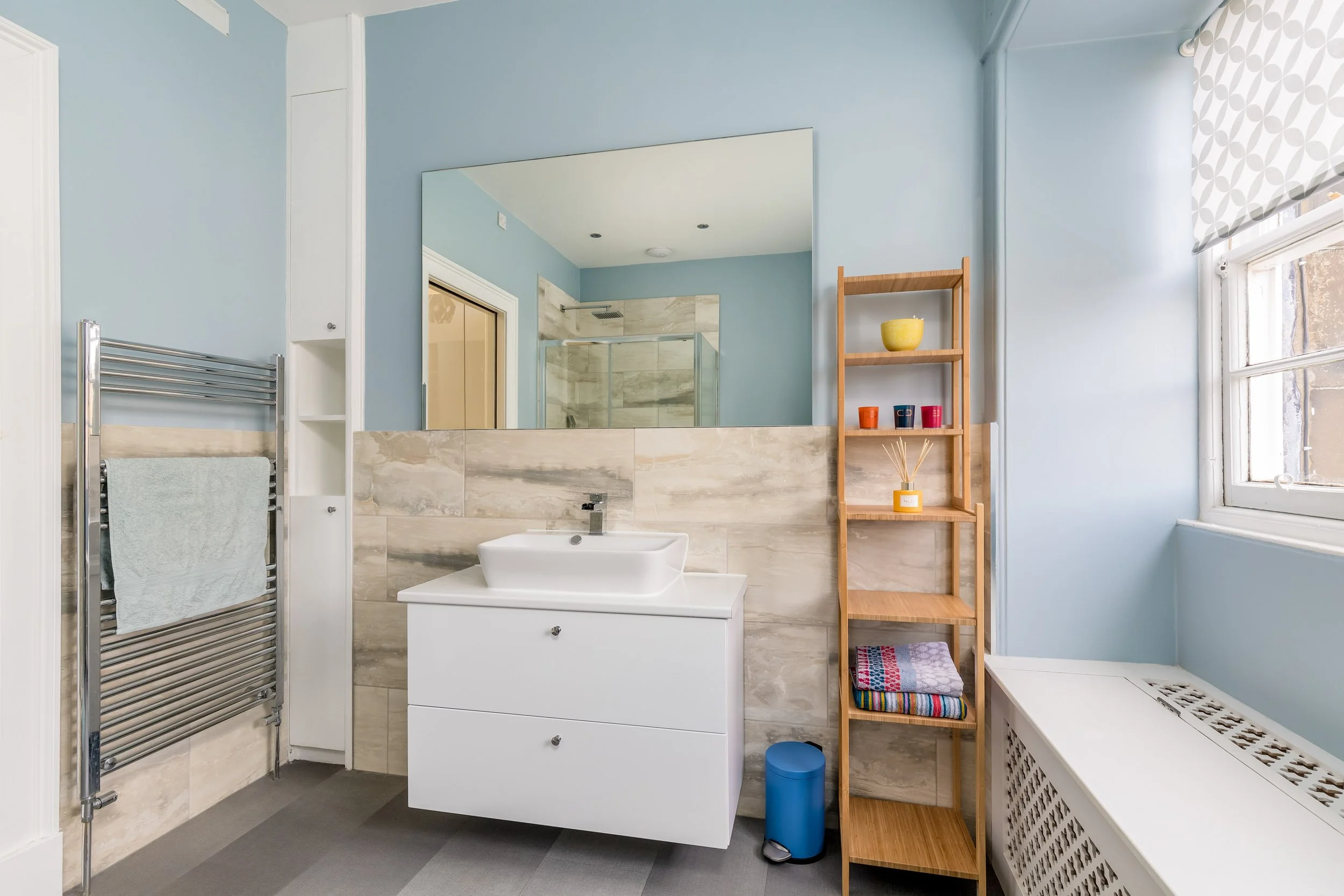 Modern bathroom with light blue walls, a large mirror, a white vanity with a vessel sink, a wooden shelving unit with colored cups and towels, a window with a patterned shade, and a tiled shower in the background.