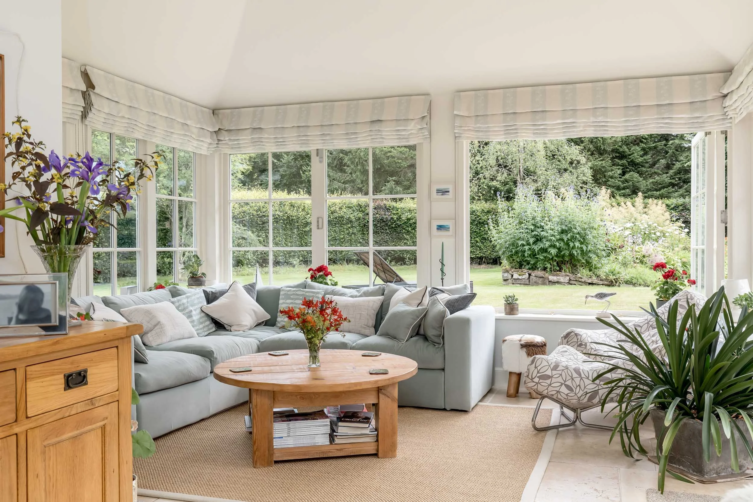 Bright living room with large windows overlooking a garden, beige carpet, light blue sectional sofa with pillows, wooden coffee table with magazines, potted plants, and floral decorations.