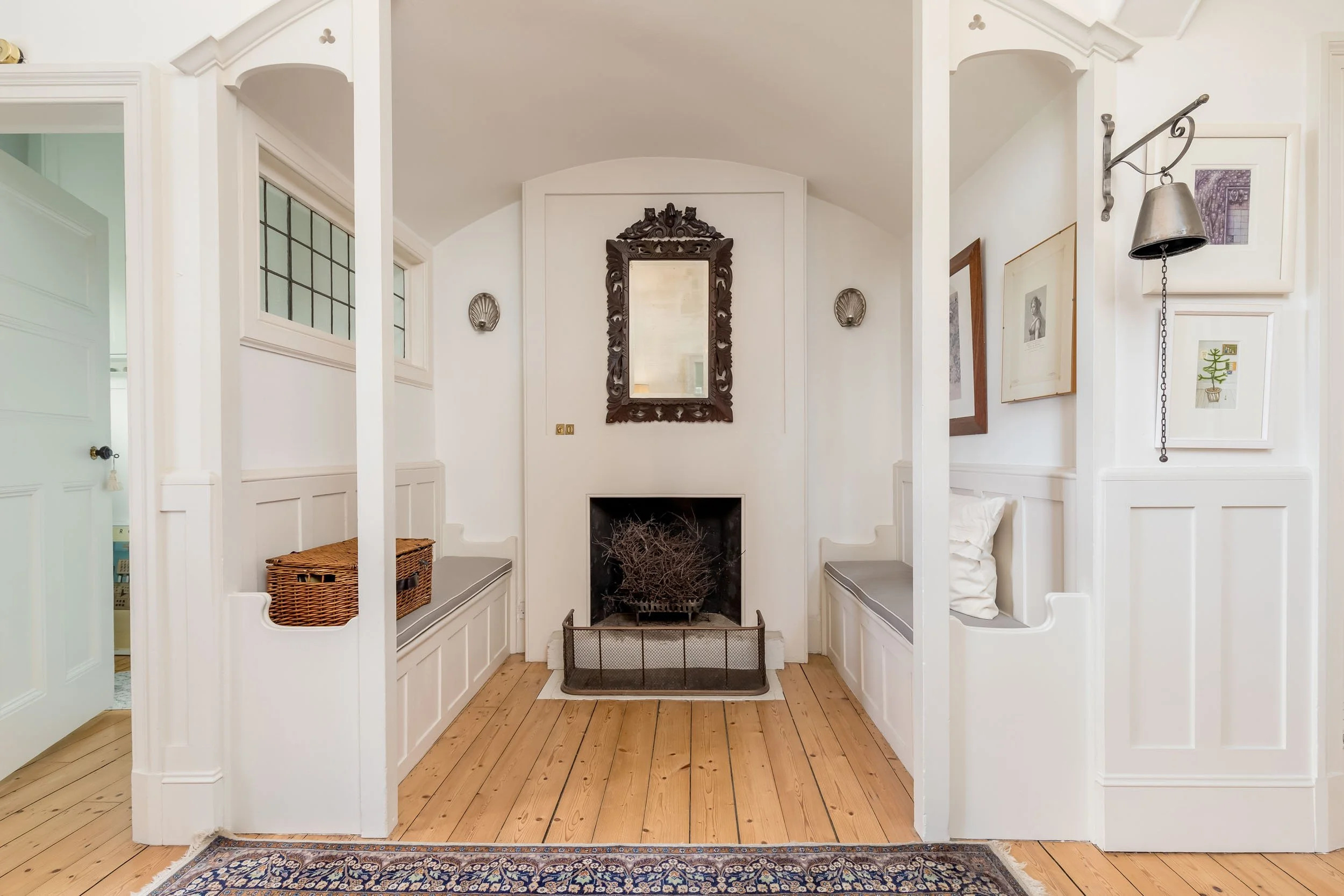 A cozy seating nook with a built-in bench on each side of a fireplace, a decorative mirror above the fireplace, framed artwork on the wall, a small rug on a wooden floor, and a vintage bell on a wall-mounted chain.