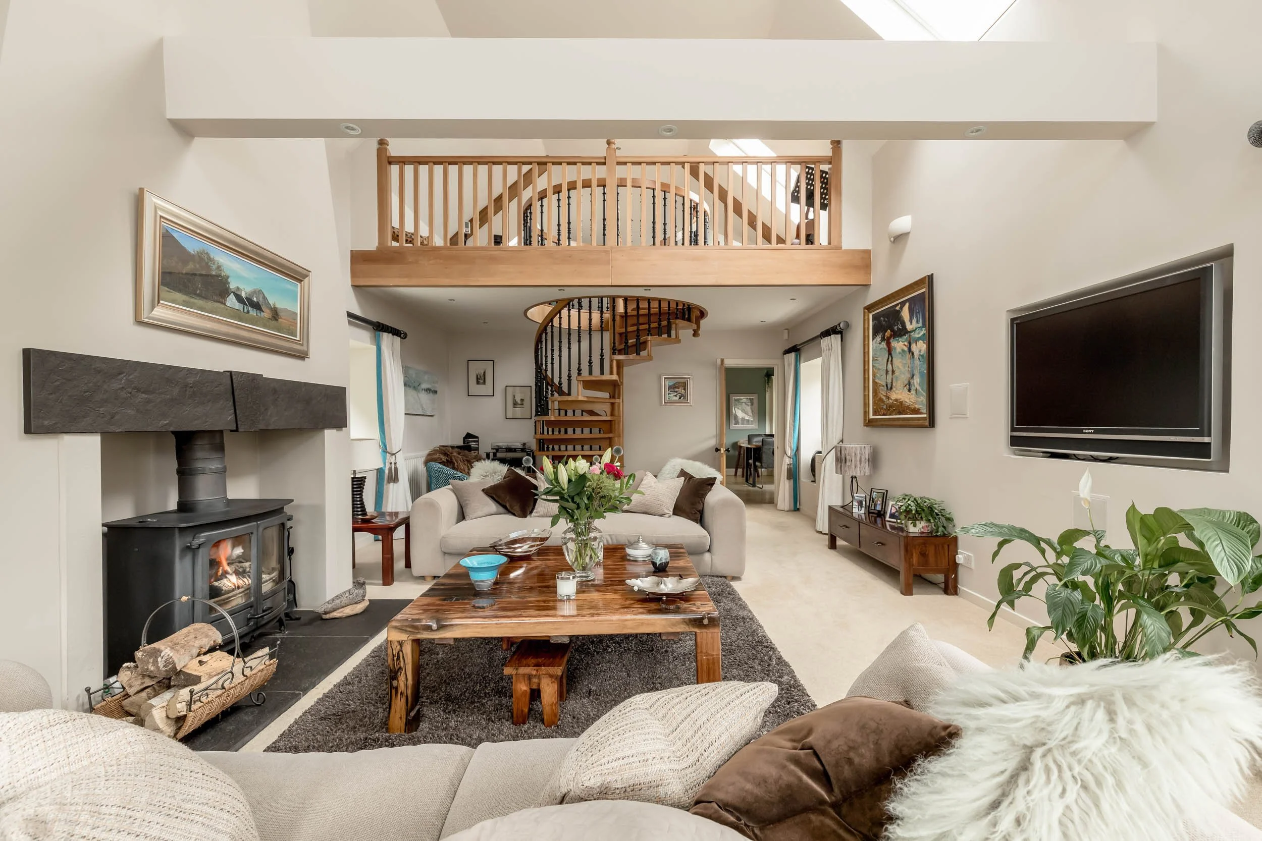 A spacious living room with a fireplace, a TV, and a second-floor balcony. The room has a beige sofa, a wooden coffee table with decorative items and a flower vase, and various artwork on the walls. There are plants near the sofa, curtains on the win