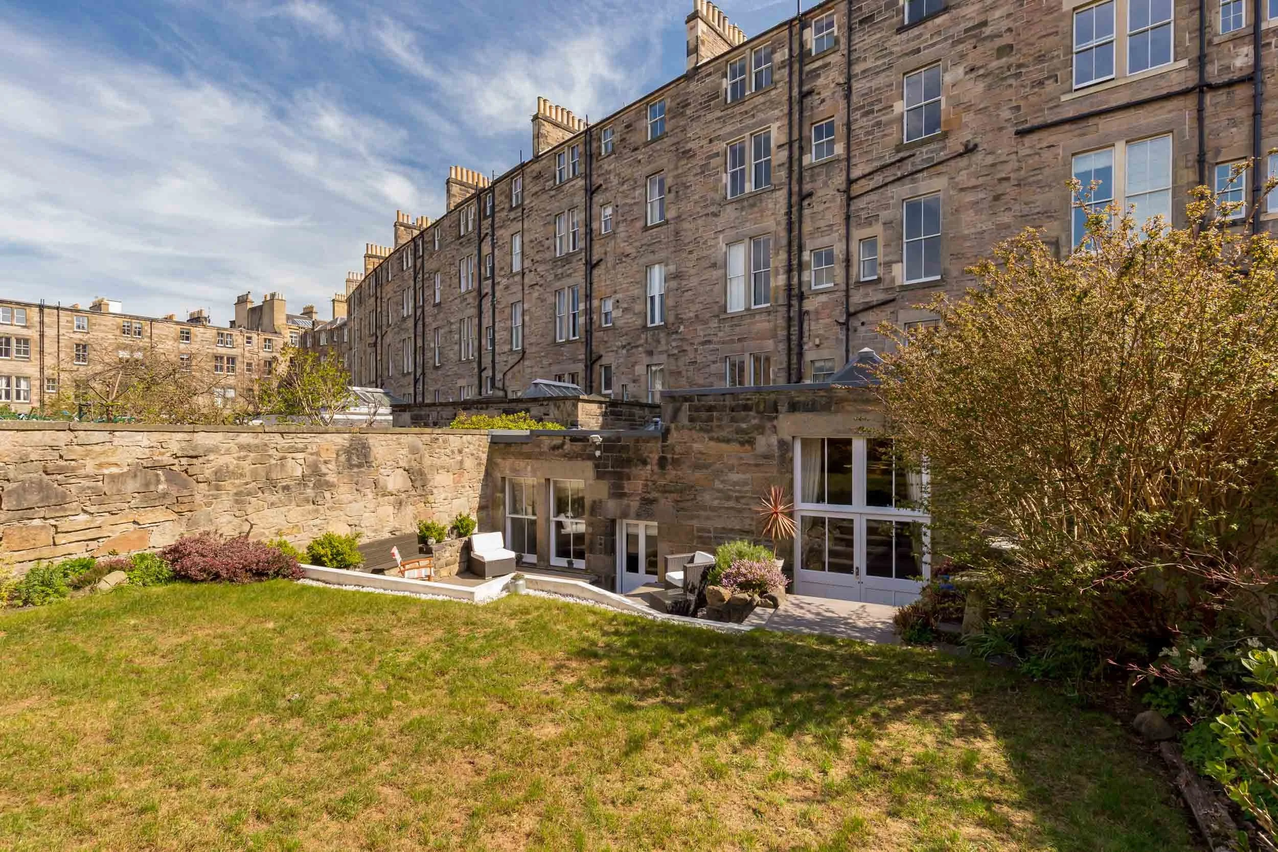A view of a garden patio area with outdoor furniture, plants, and a grassy lawn, surrounded by a stone wall and adjacent to a multi-story stone apartment building under a partly cloudy sky.