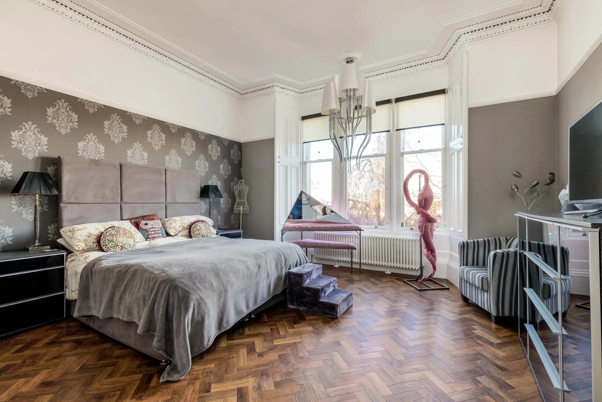 A spacious bedroom with a large bed featuring a gray headboard, patterned pillows, and a gray blanket. The room has dark and light gray walls, with one wall decorated with a floral wallpaper. There are two black nightstands with lamps, a pyramidal de