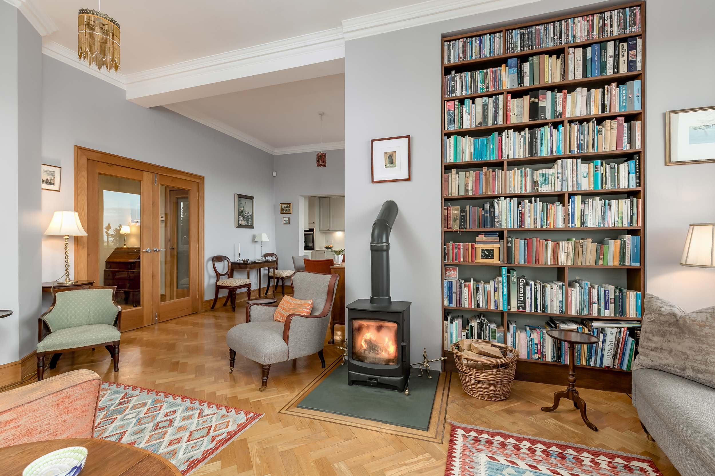 Living room with a wood stove, a large bookshelf filled with books, armchairs, a small table, lamps, a basket, and hardwood floors.