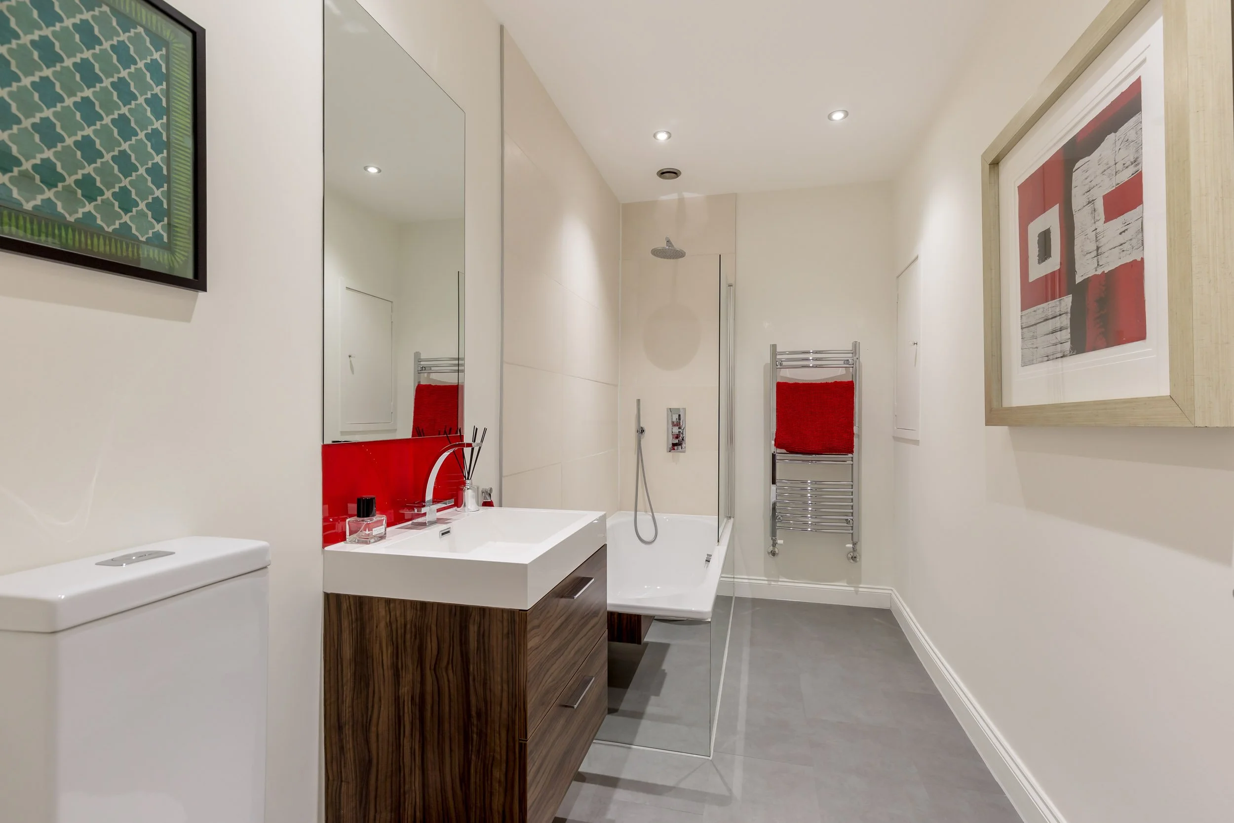 Modern bathroom with a white sink, wooden vanity, large mirror, bathtub with a glass partition, and a heated towel rack with red towels. There are framed artworks on the walls and recessed ceiling lights.
