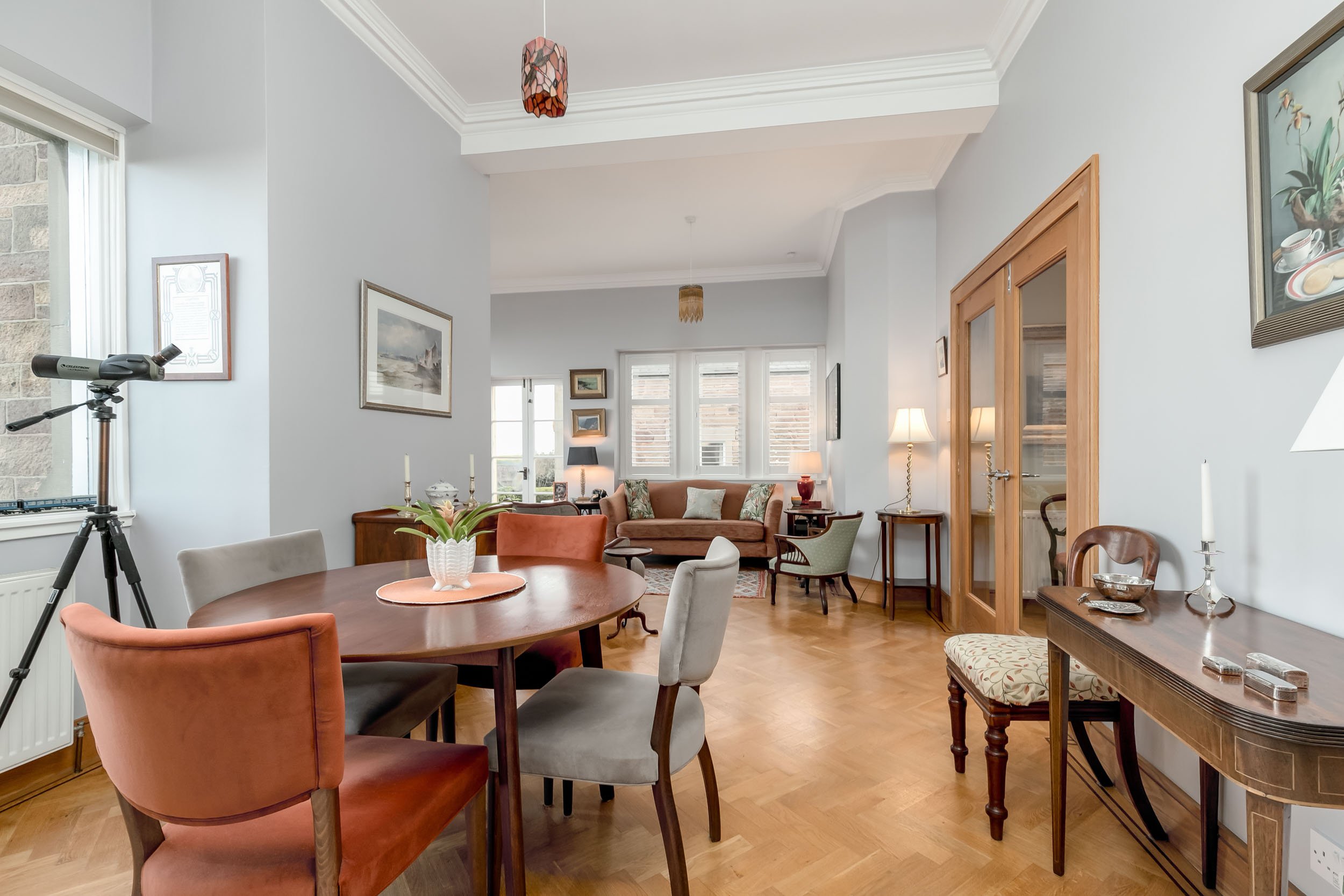 A spacious living room with a round dining table surrounded by six chairs, a telescope near the window, a sofa set with cushions, and various lamps and framed pictures on the walls.