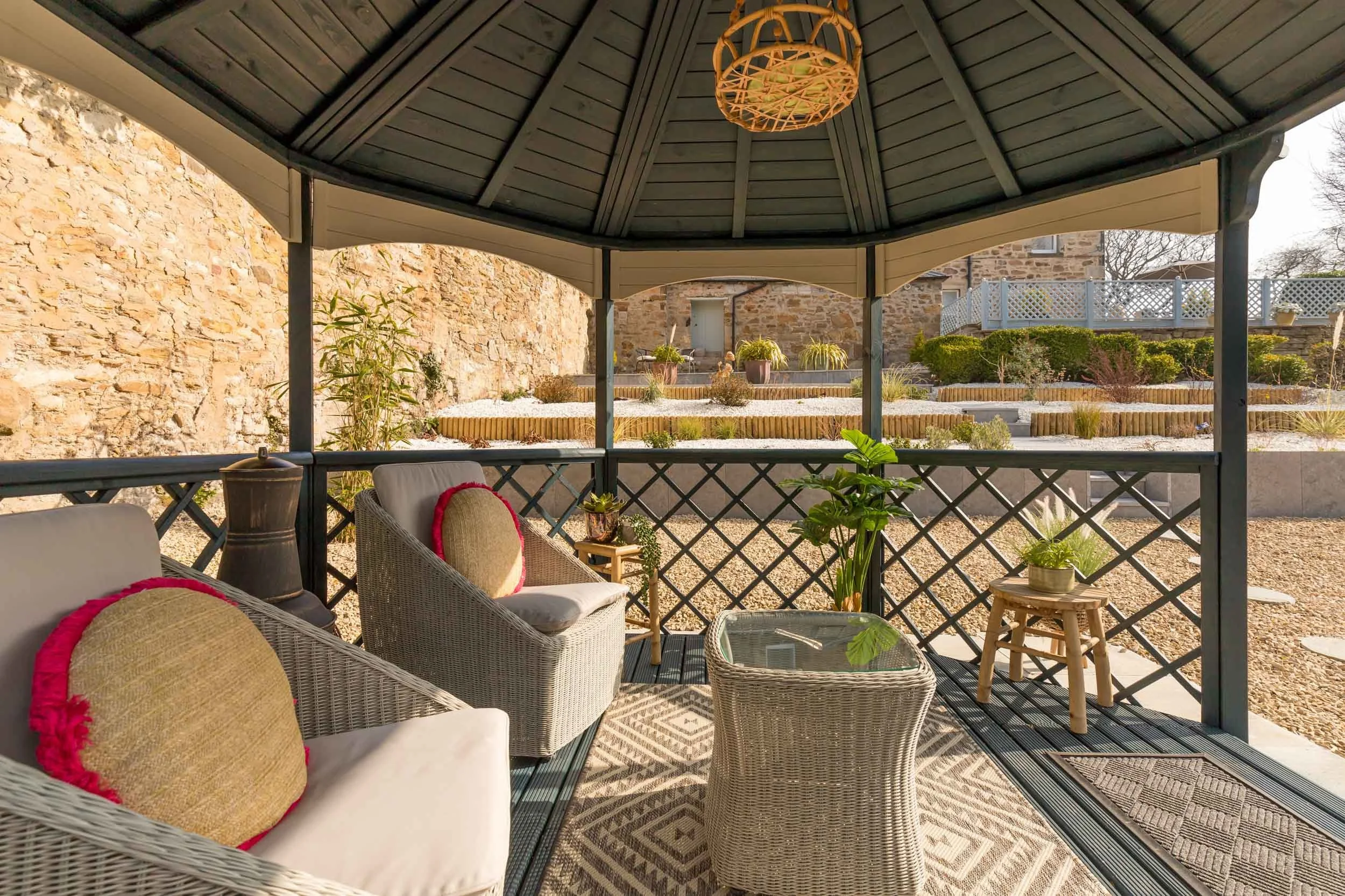 Outdoor patio with wicker furniture, houseplants, and a rug beneath a dark green gazebo with a woven light fixture, overlooking a landscaped yard with plants and stone walls.