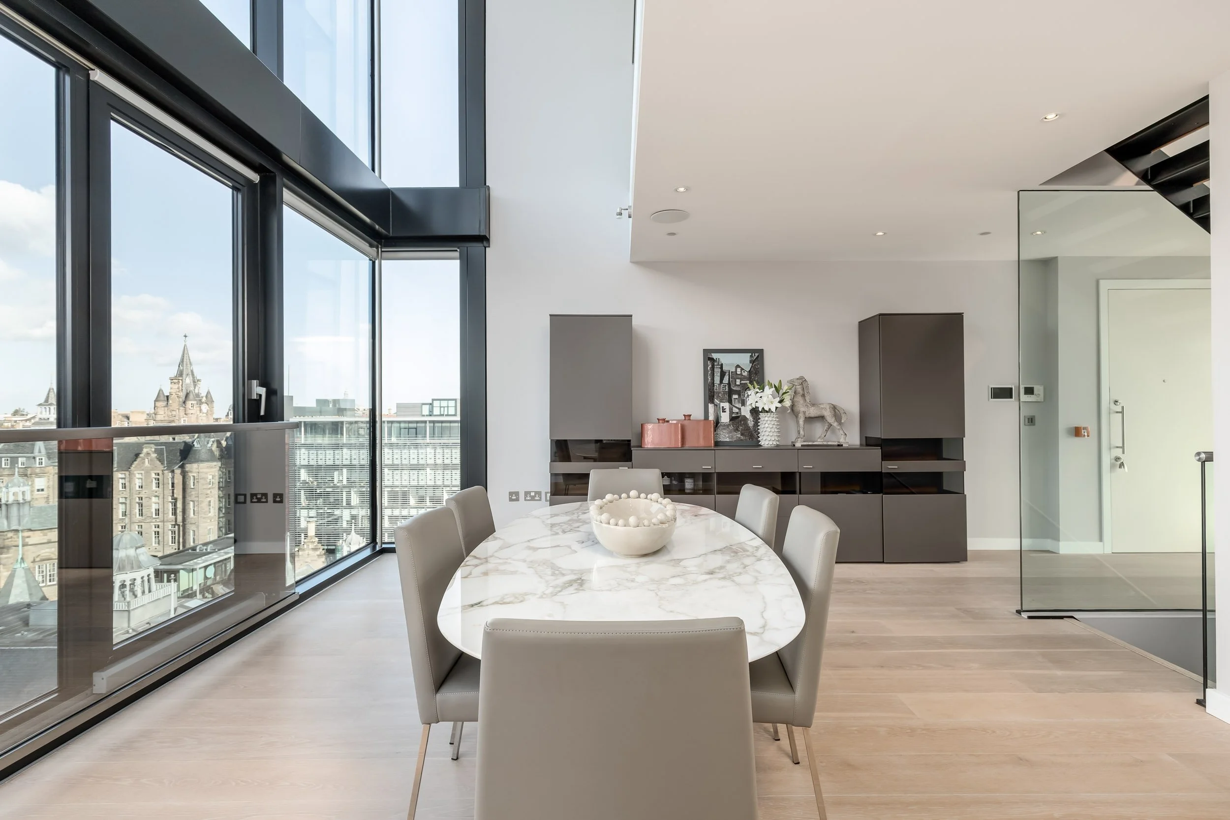 Modern dining room with a marble-top oval table and six beige chairs, large floor-to-ceiling windows with city view, white walls, grey cabinets, decorative items, and a glass wall separating the staircase area.