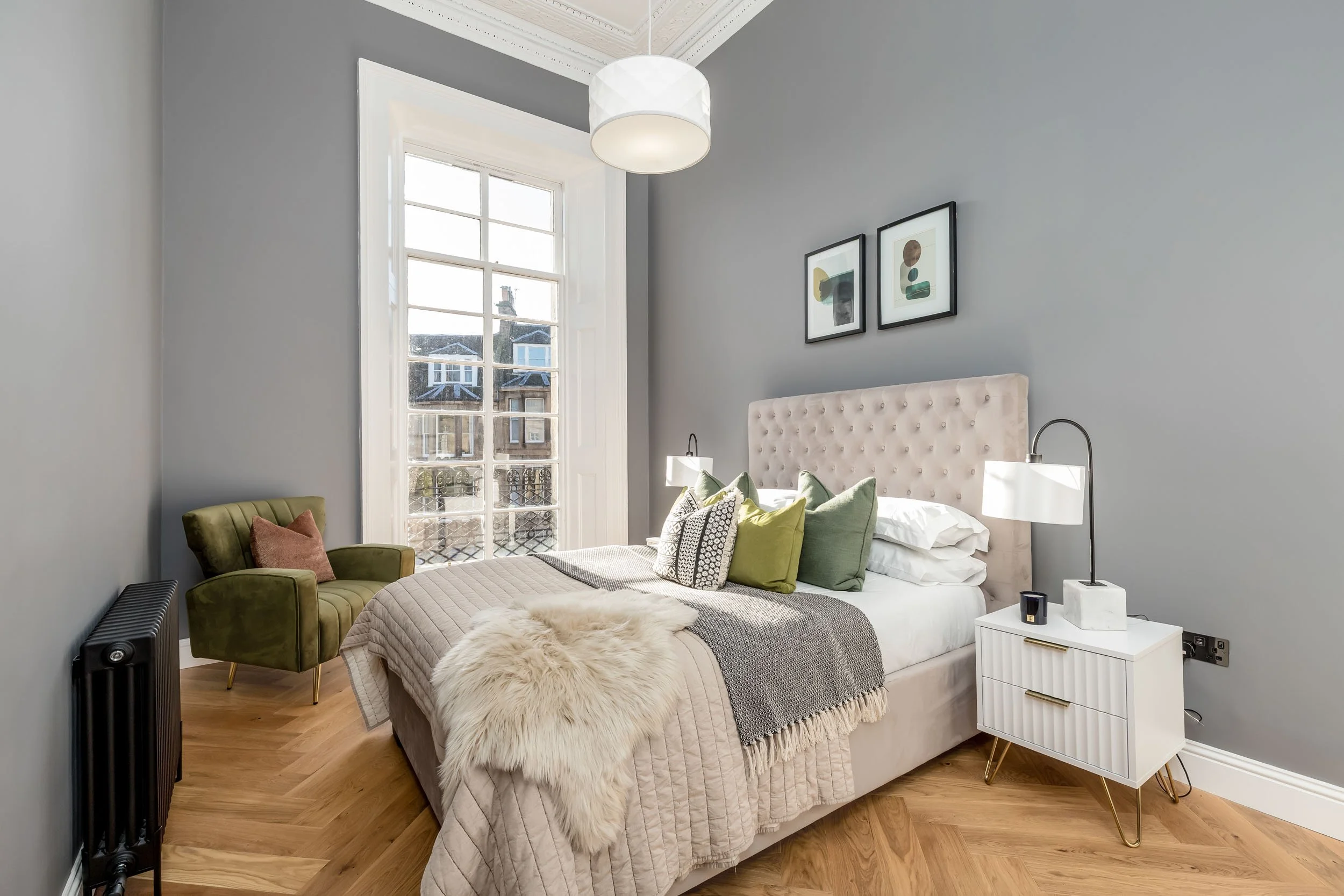 A modern bedroom featuring a large beige tufted headboard, a white side table with gold accents, and a collection of green, black, and white decorative pillows on the bed. There is a white fur throw on the bed, a green armchair with a rust-colored pi