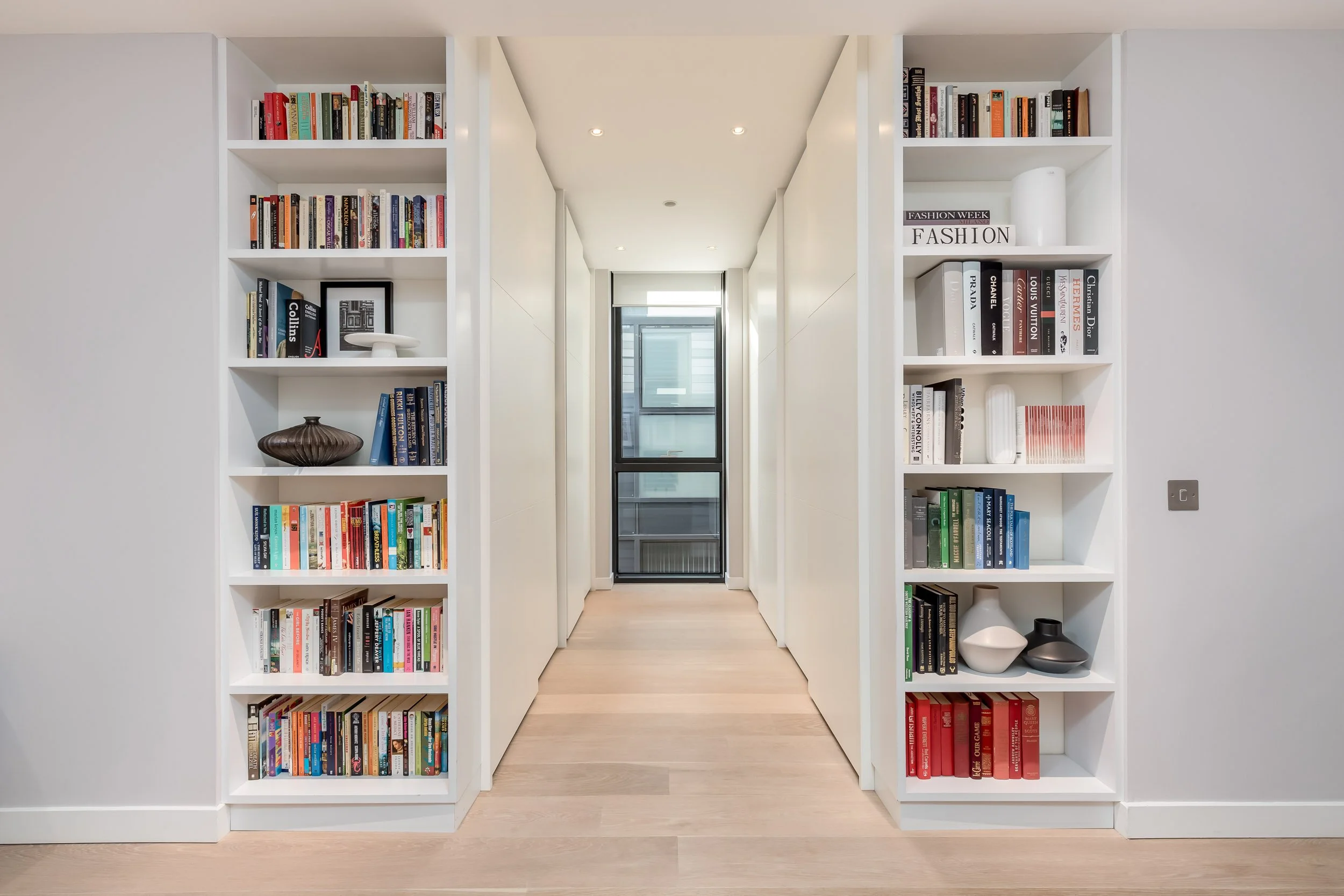 A modern hallway with white built-in bookshelves on both sides, filled with books and decorative objects, leading to a glass door at the end.