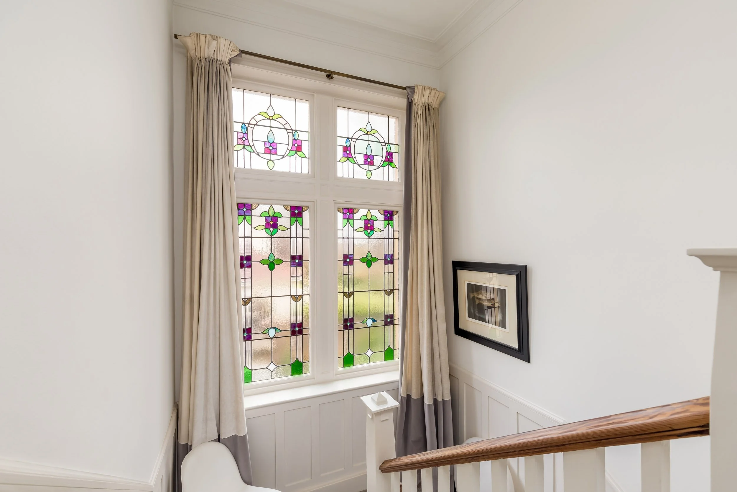 Interior view of a staircase landing with a large stained glass window, cream curtains, and a framed picture on the wall.