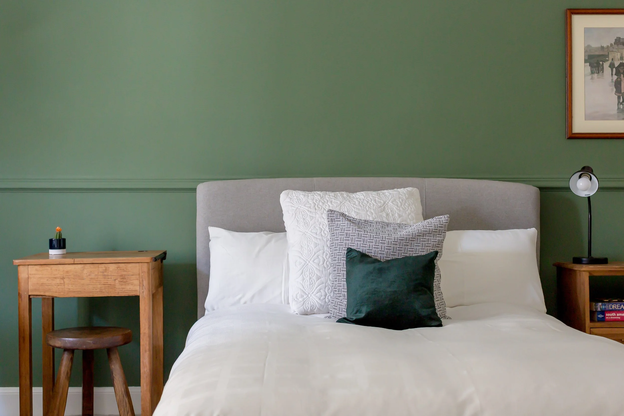 Bedroom with green wall, gray headboard, white bedding, decorative pillows, wooden side tables, lamp, framed artwork.