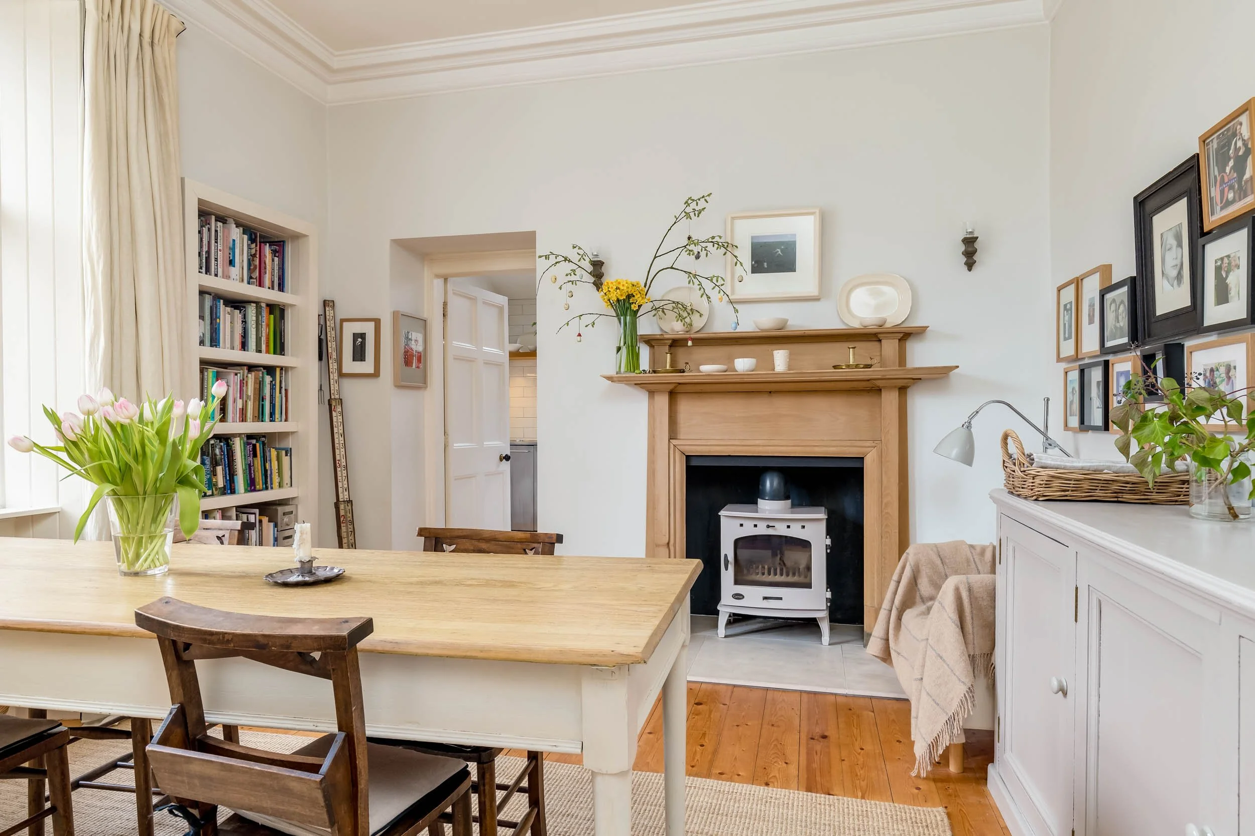 Living room with a dining table, a vase of pink tulips, a white cabinet with plants and framed photos, a fireplace with a wood mantel decorated with flowers, ceramics, and art, a wood floor, and a doorway leading to the kitchen.