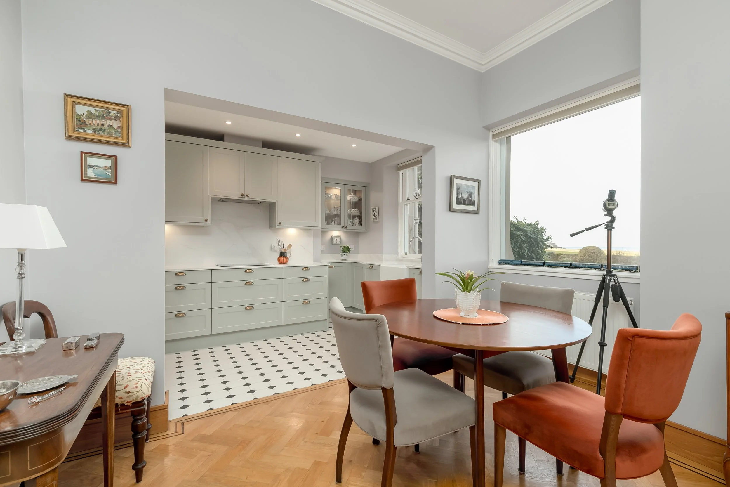 A dining room with a wooden table, six upholstered chairs, a potted plant in the center, and a large window with a view outside. A camera on a tripod is set up near the window. In the background, a kitchen with white cabinets, a marble backsplash, an