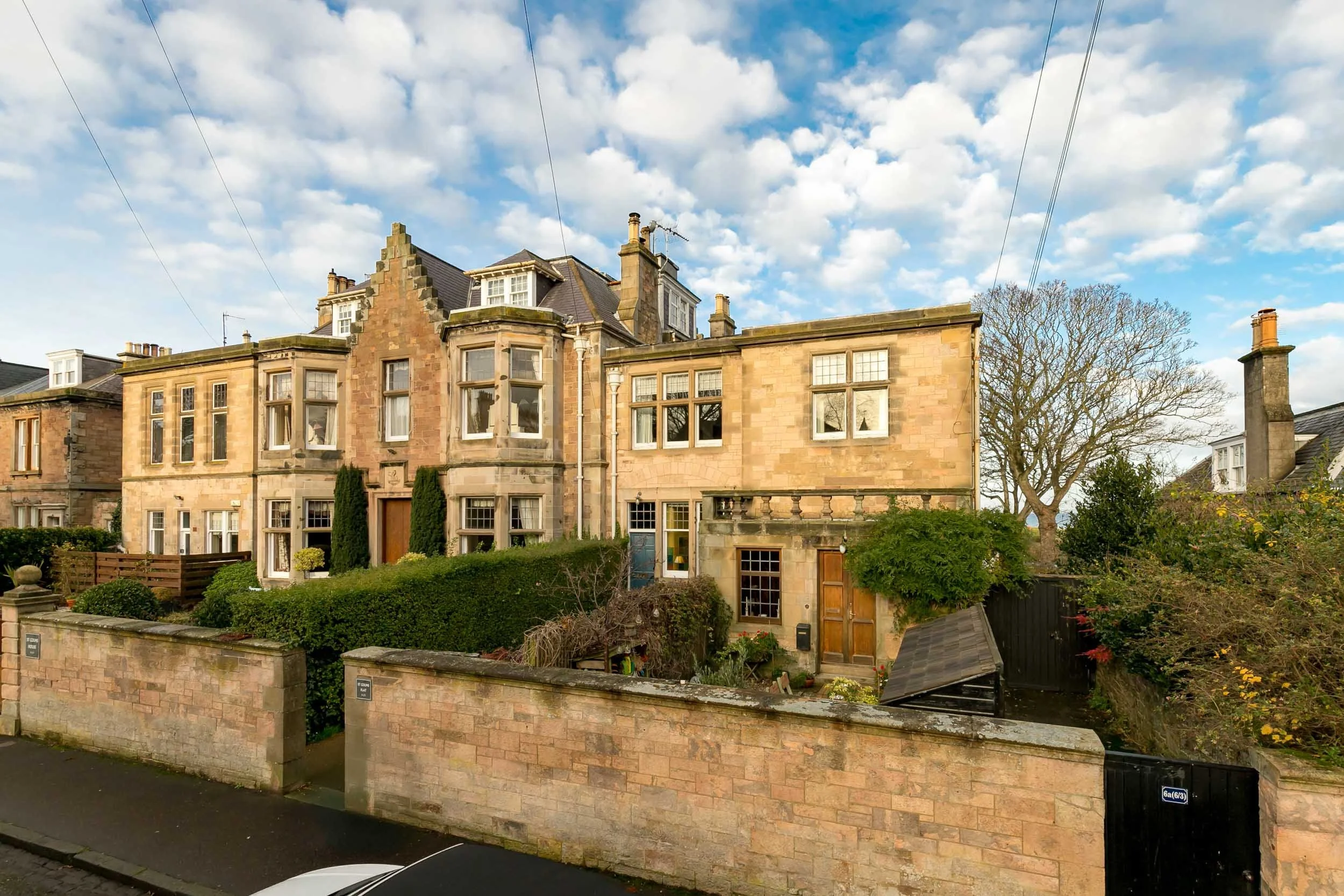 A large, historic stone building with tall windows and a gabled roof, surrounded by a brick wall and greenery, under a partly cloudy sky.