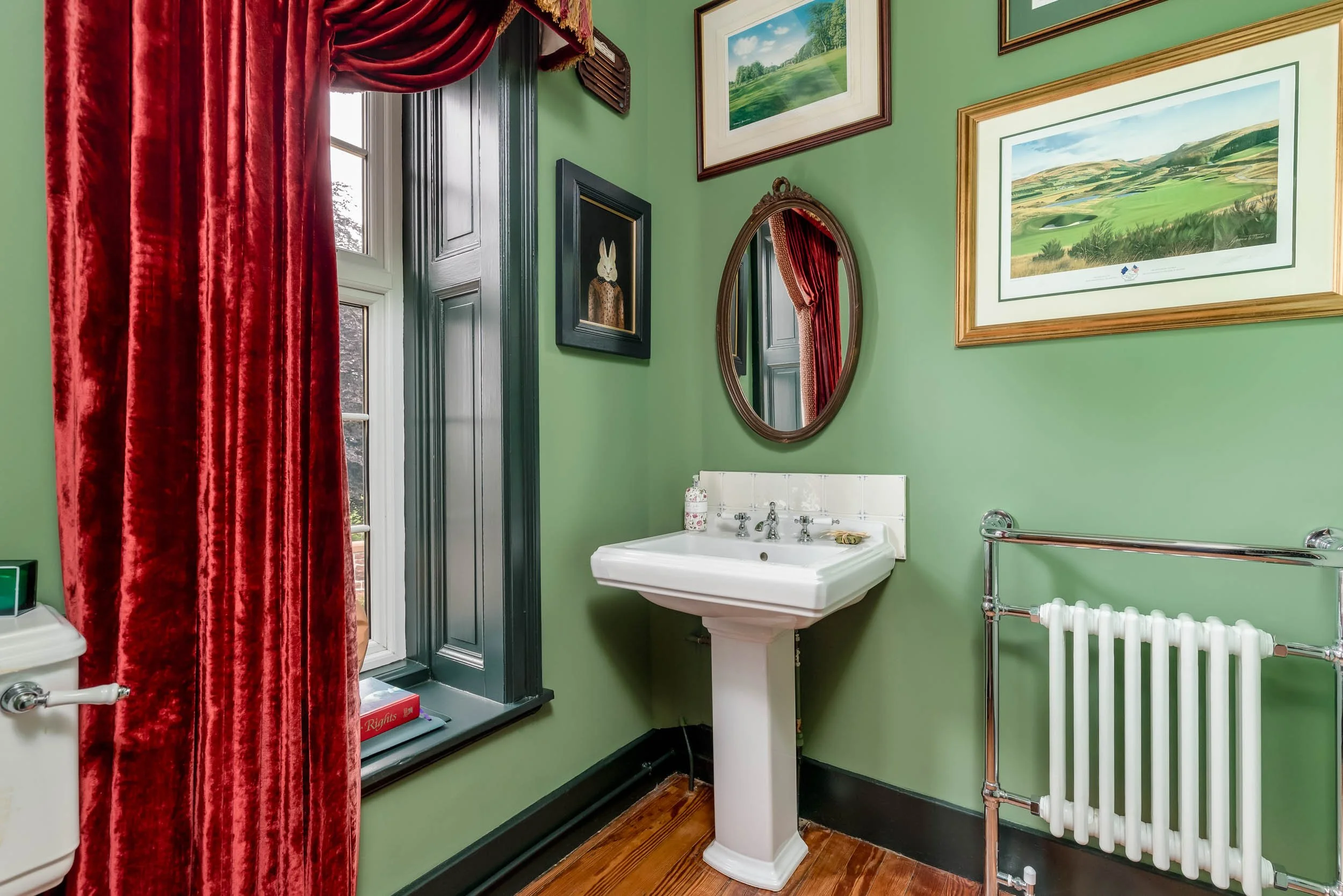 A vintage bathroom featuring green walls, a white pedestal sink, framed landscape pictures, a small oval mirror, a radiator, and a window with red velvet curtains.