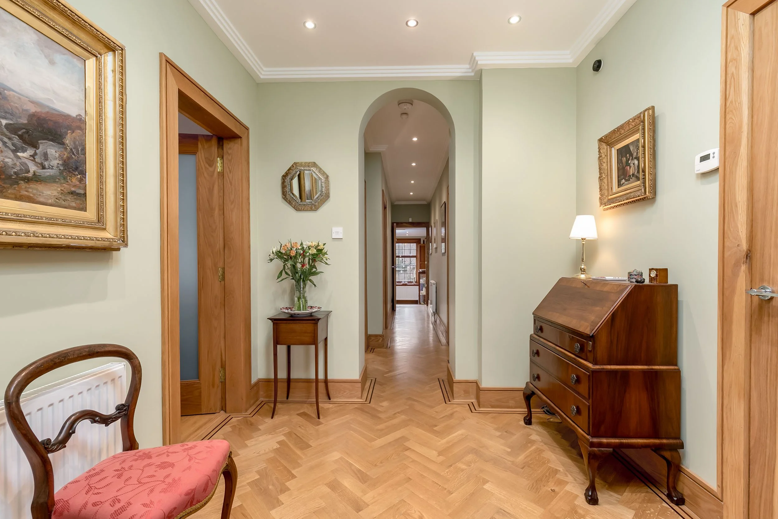 Interior hallway with wooden flooring, light green walls, and classic furniture, including a pink upholstered chair and a wooden secretary desk, with decorative paintings and a small table with flowers.