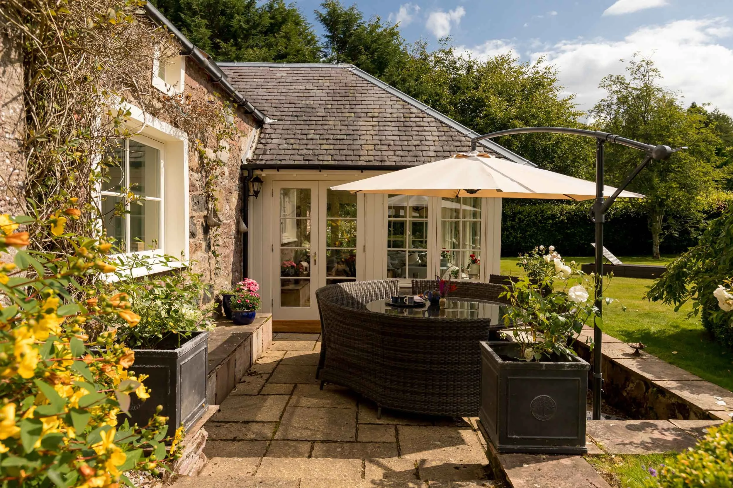 Outdoor patio area with round wicker table, chairs, large white umbrella, potted flowers, stone pathway, and a stone house with glass door and window, surrounded by a lush green garden.