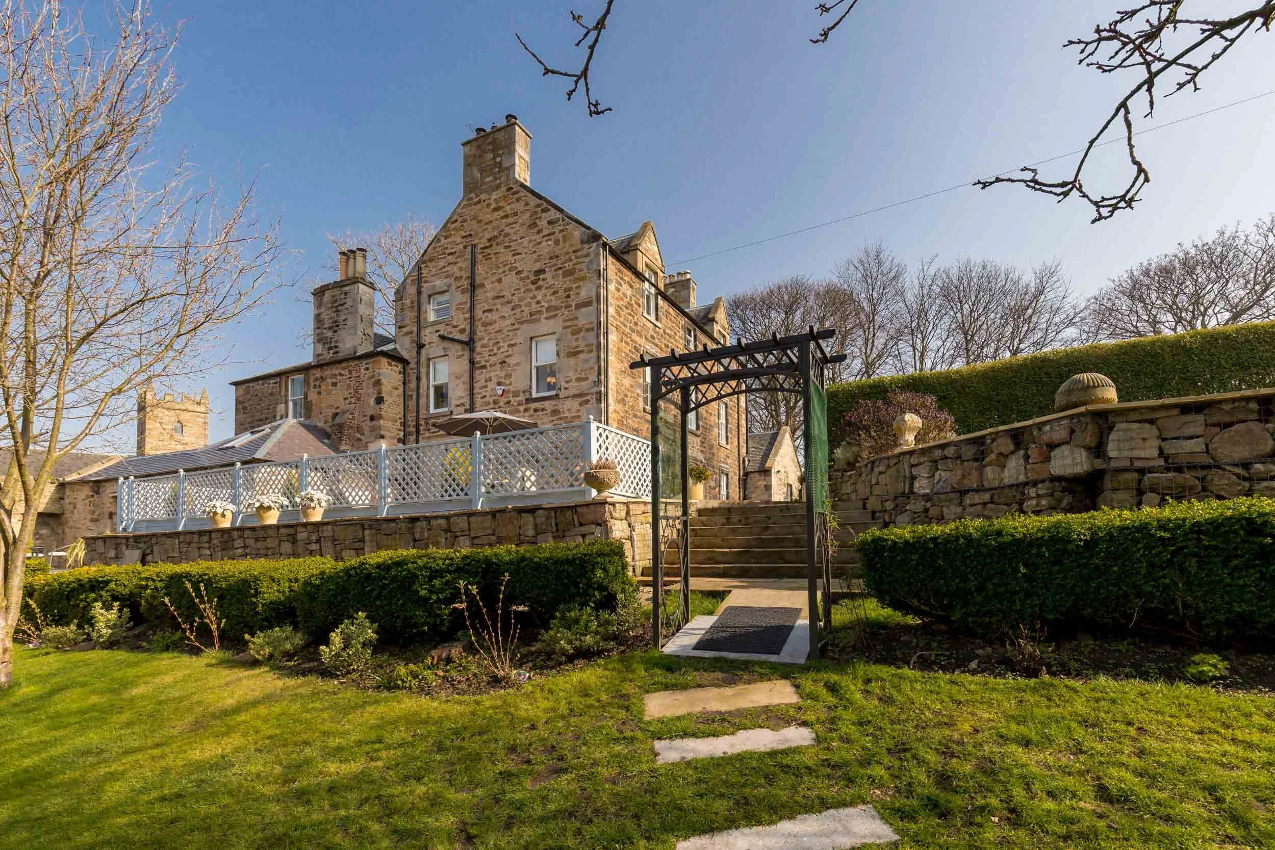 A historic stone house with a garden, stone steps, and a white picket fence, under a clear blue sky.