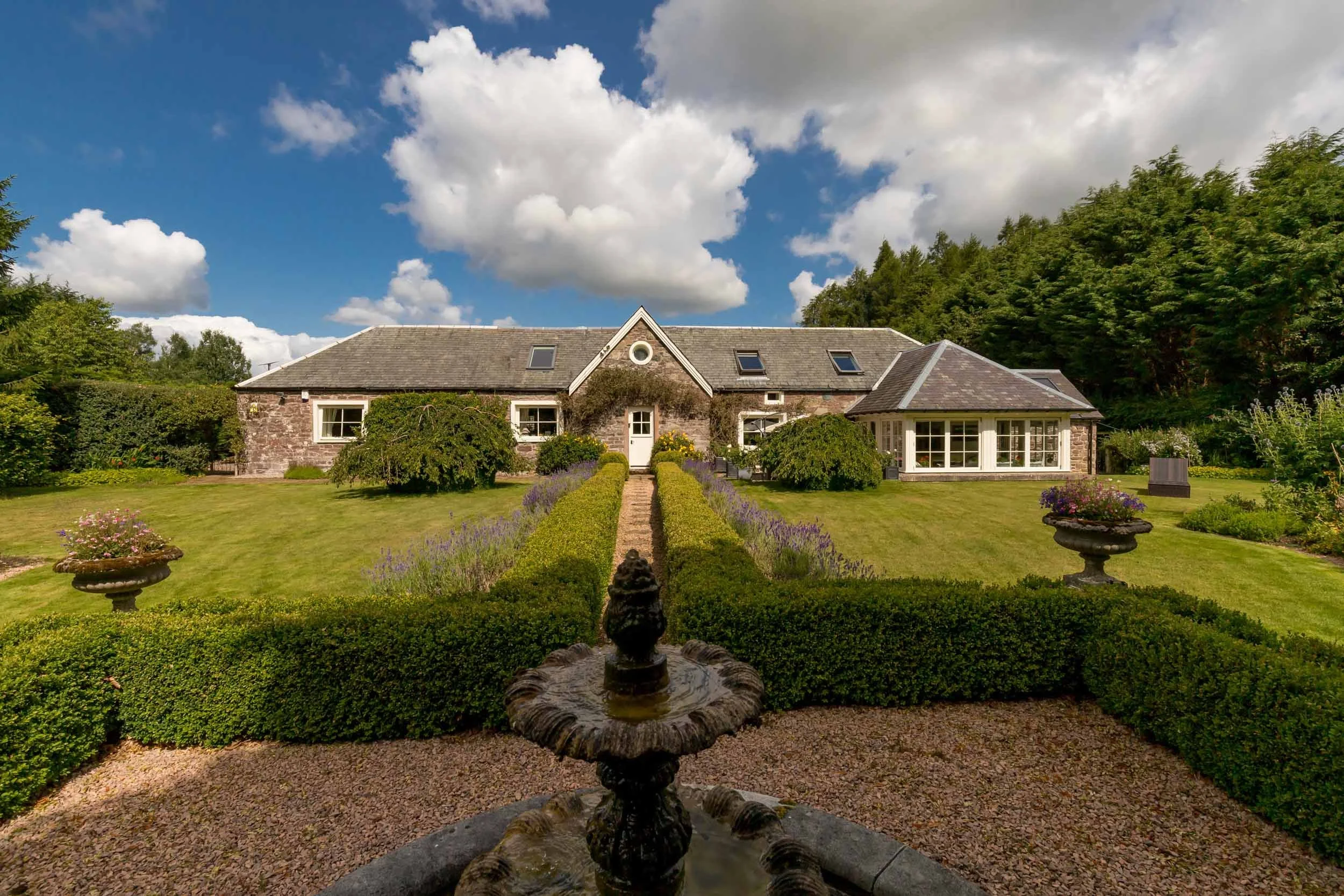 A large stone house with a gabled roof, multiple windows, and a white door, surrounded by a well-maintained garden with trimmed bushes, flowering plants, and two large trees on either side, under a partly cloudy sky.