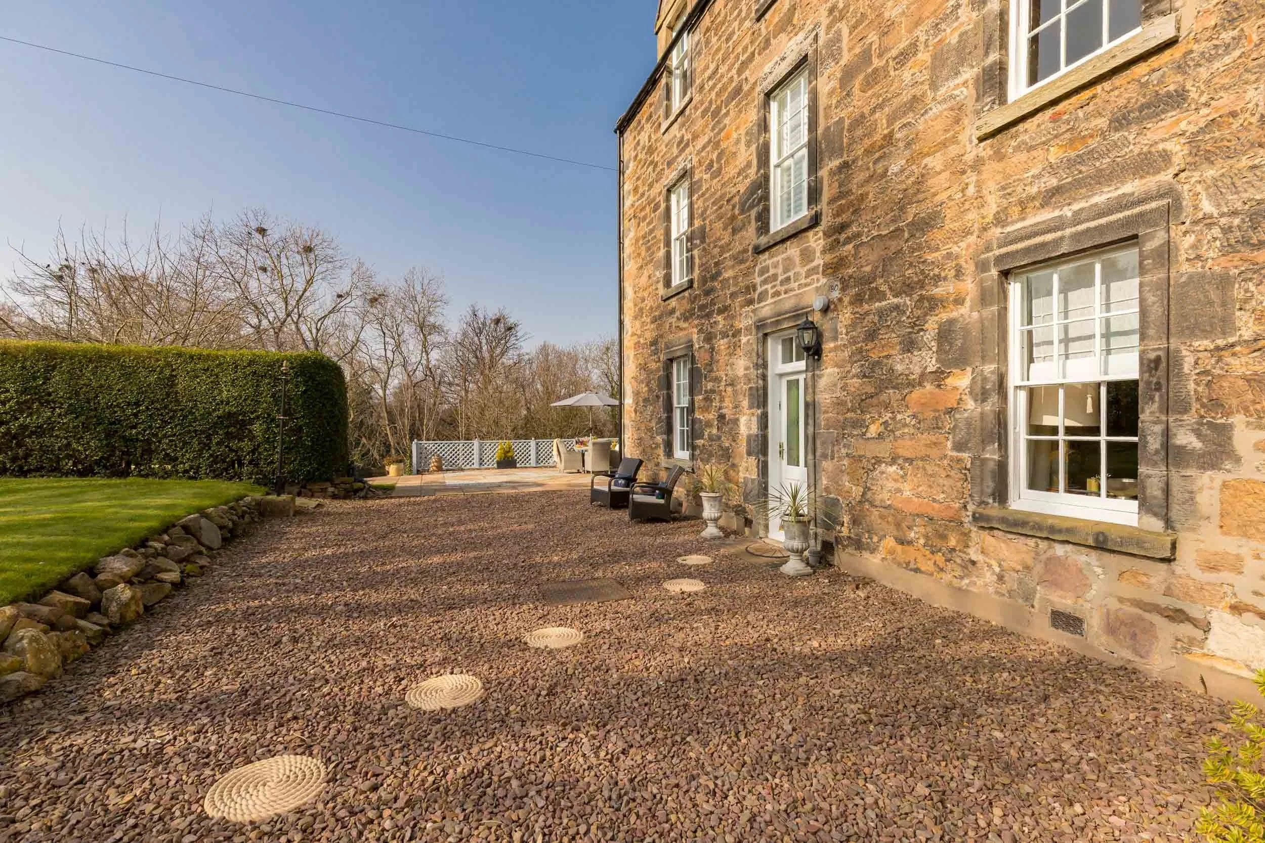Stone house exterior with gravel patio, white-framed windows, and black benches, overlooking a landscaped yard with green bushes and trees in the background.
